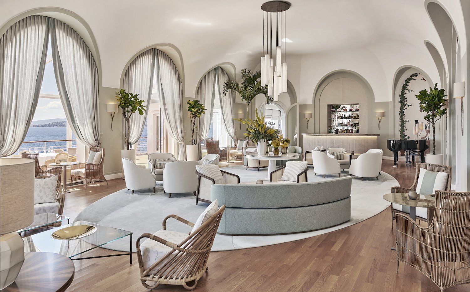 Intérieur d'un salon élégant avec des grands fenêtres offrant une vue sur la mer, mobilier blanc, fauteuils, plants d'intérieur, un piano noir, et un bar en arrière-plan.