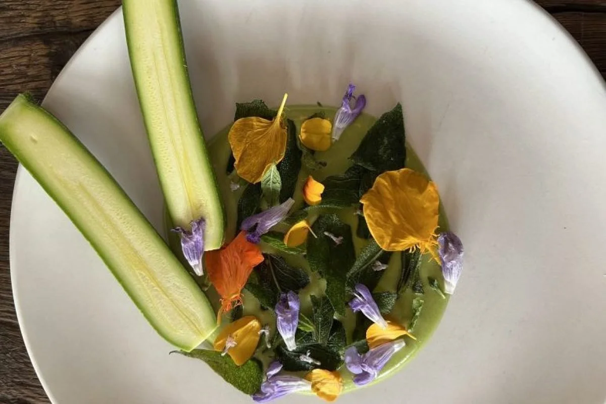 Une assiette blanche contenant de l'eau avec des feuilles et des fleurs comestibles de différentes couleurs, ainsi que deux bâtonnets de céleri coupés en deux, aux extrémités vertes.