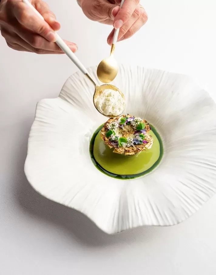 Une assiette blanche avec un plat gastronomique, garnie de fleurs comestibles, avec une sauce verte, et deux mains versant une sauce ou un crémeux doré sur le plat.