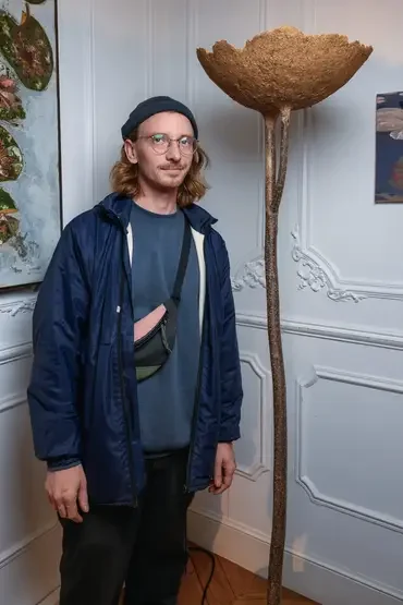 Un homme avec des cheveux longs, lunettes, chapeau noir, portant un sac à l'épaule et une veste bleue, se tient à côté d'une sculpture en forme de champignon géant dans une pièce avec décor blanc.