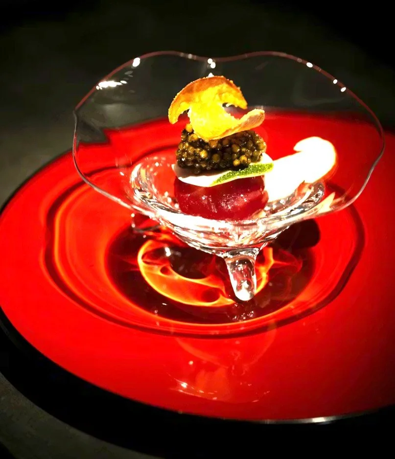 Un plat gastronomique dans un verre en cristal, avec des ingrédients comme de la caviar, une tranche de citron vert, et une coquille d'œuf, présenté sur une assiette rouge