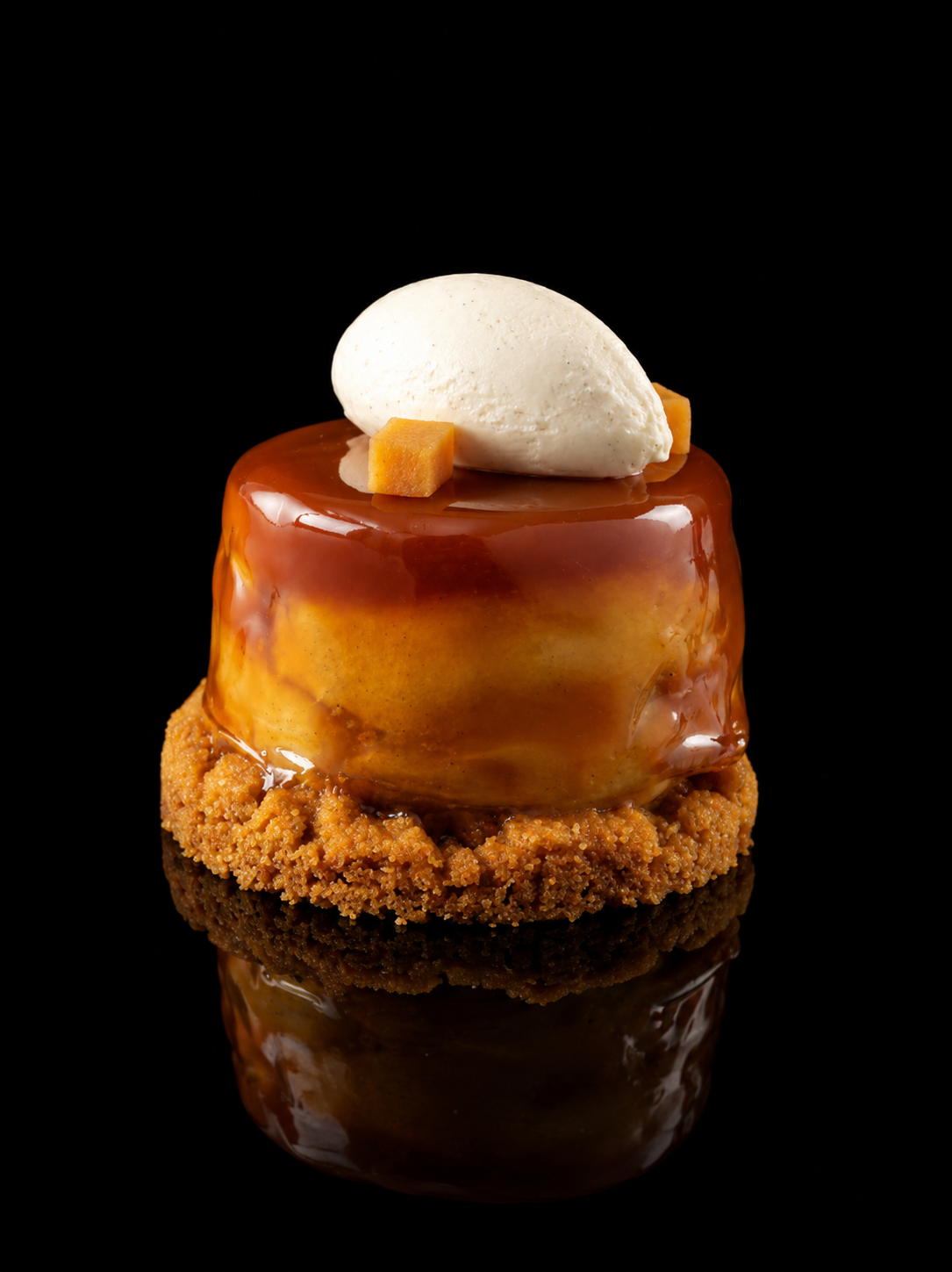 Gâteau pâtissier avec une couche de caramel brillant, décoré avec une boule de crème ou glace à la vanille et deux petits cubes de mangue, posé sur une base croquante.