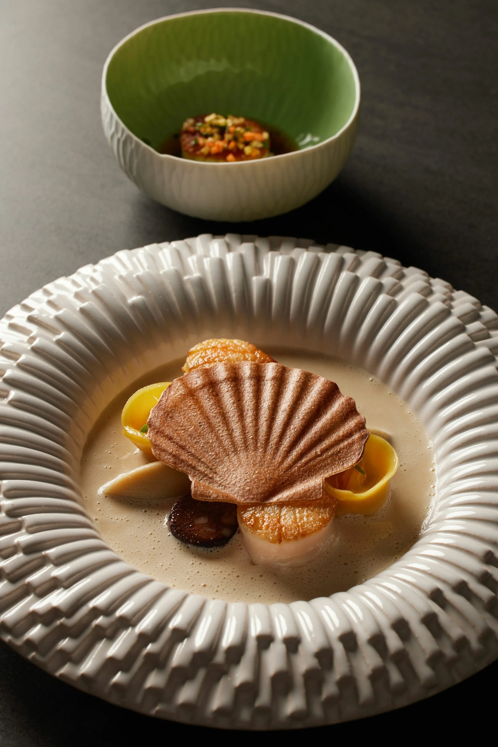 Un plat gastronomique avec des coquilles Saint-Jacques, accompagné d'une sauce ou d'une crème, et une petite assiette verte avec un autre plat ou une garniture en arrière-plan.