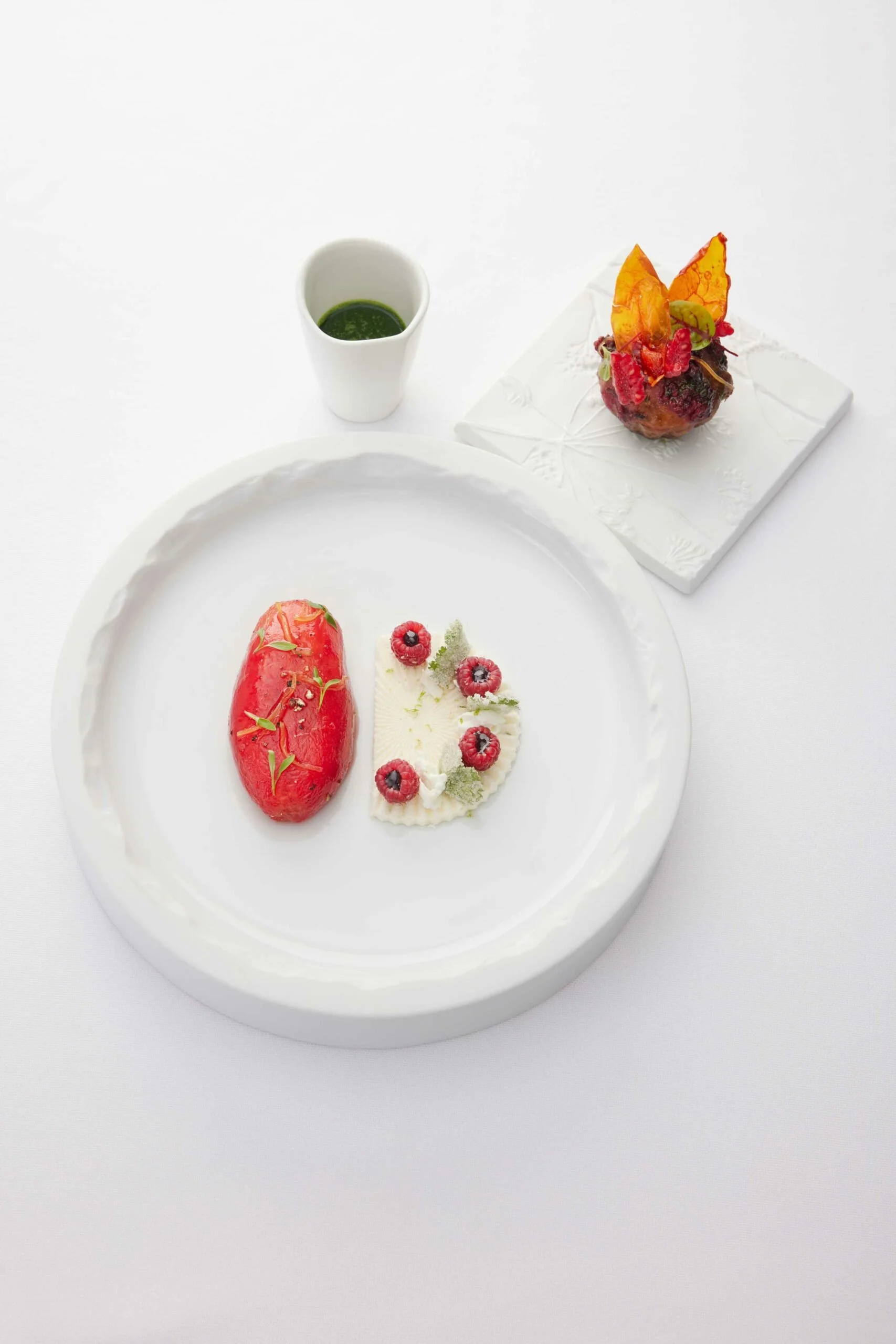 Un plat de dessert élégant avec une pêche ou un pamplemousse rouge, un petit gâteau décoré de framboises, une sauce ou purée rouge dans un petit bol, et une autre petite pâtisserie décorée de fruits rouges et feuilles vertes, le tout présenté sur une