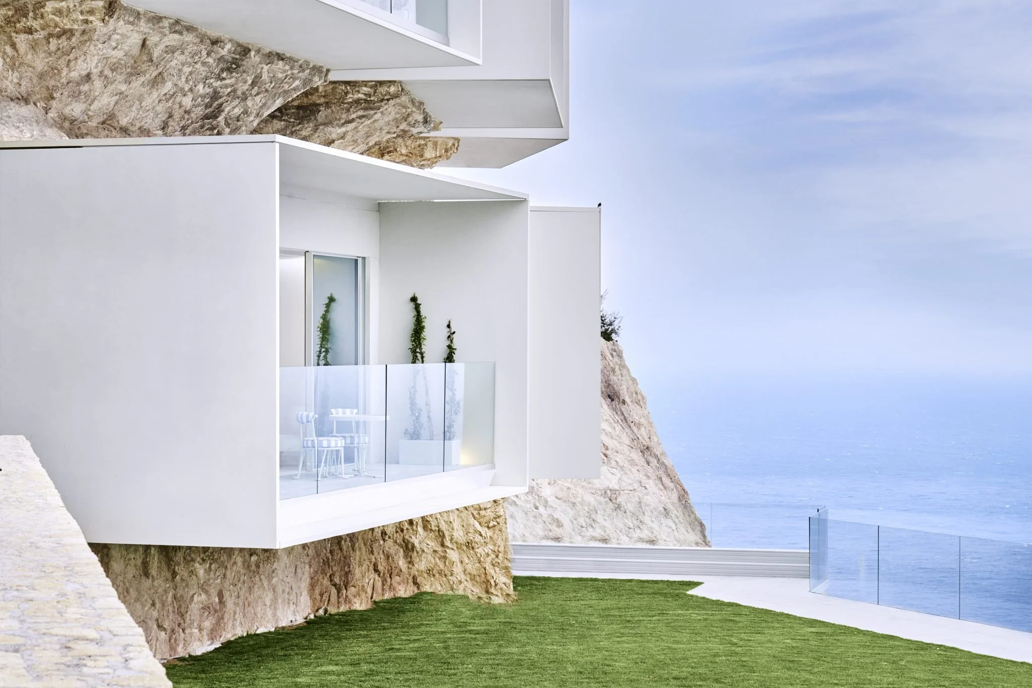 Une maison moderne blanche construite sur une falaise au bord de la mer avec un balcon en verre et un espace vert devant.