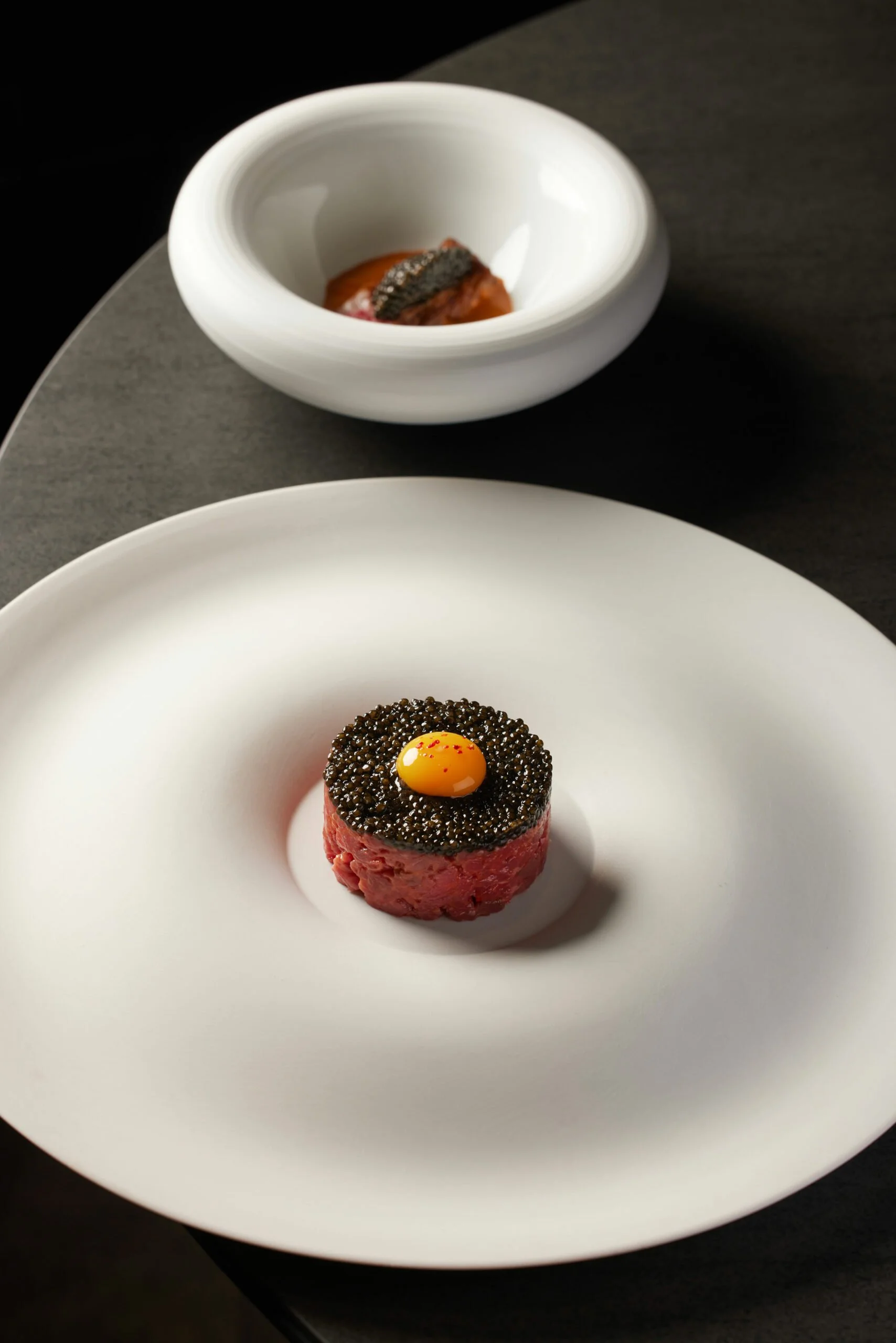 Un plat de viande rouge servie avec une couche de caviar noir, garnie d'un œuf de caille jaune sur le dessus, présenté sur une assiette blanche.