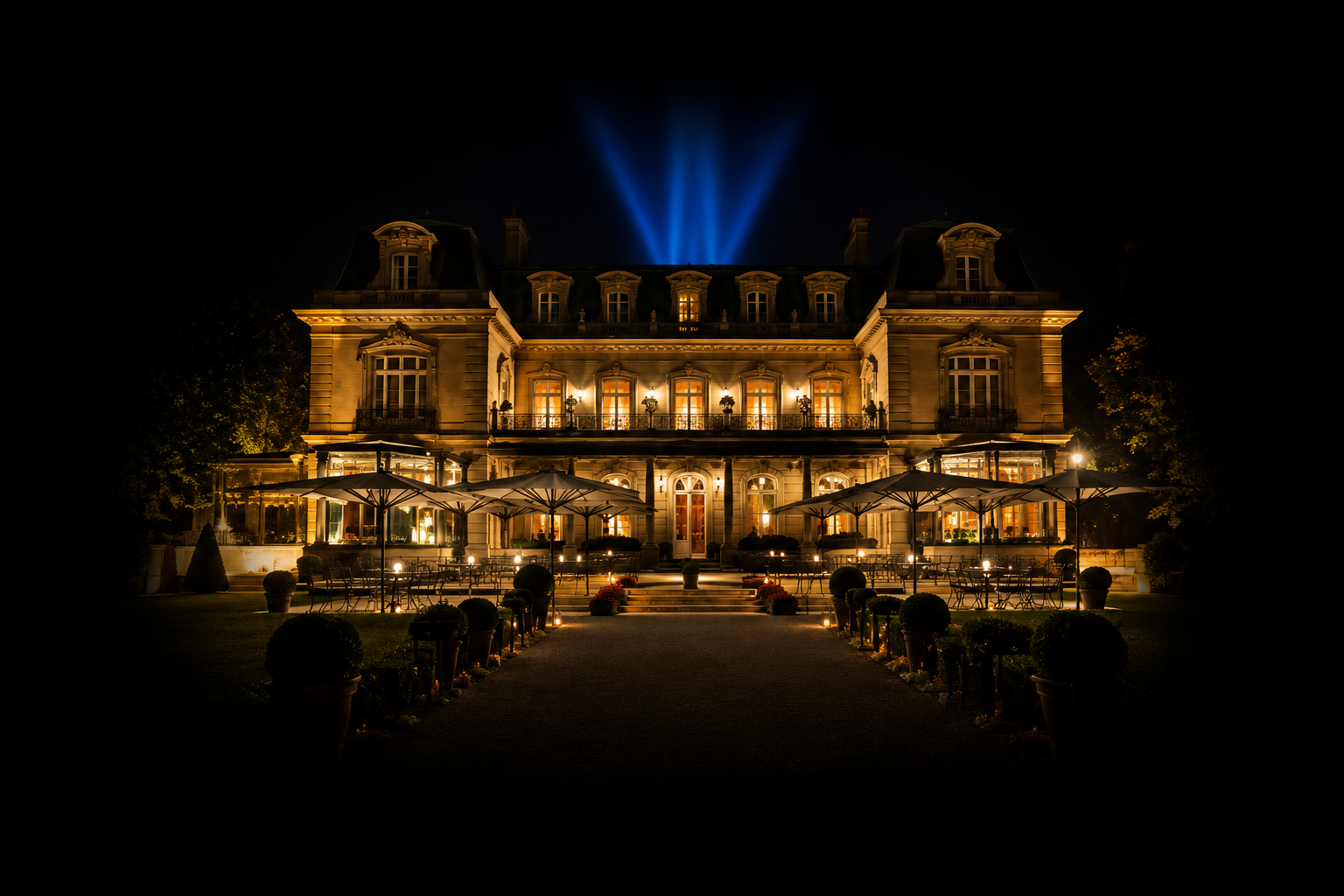 Château éclairé la nuit avec des lumières chaudes, terrasse avec parasols et mobilier, avec un chemin bordé de plantes en pot et de bougies