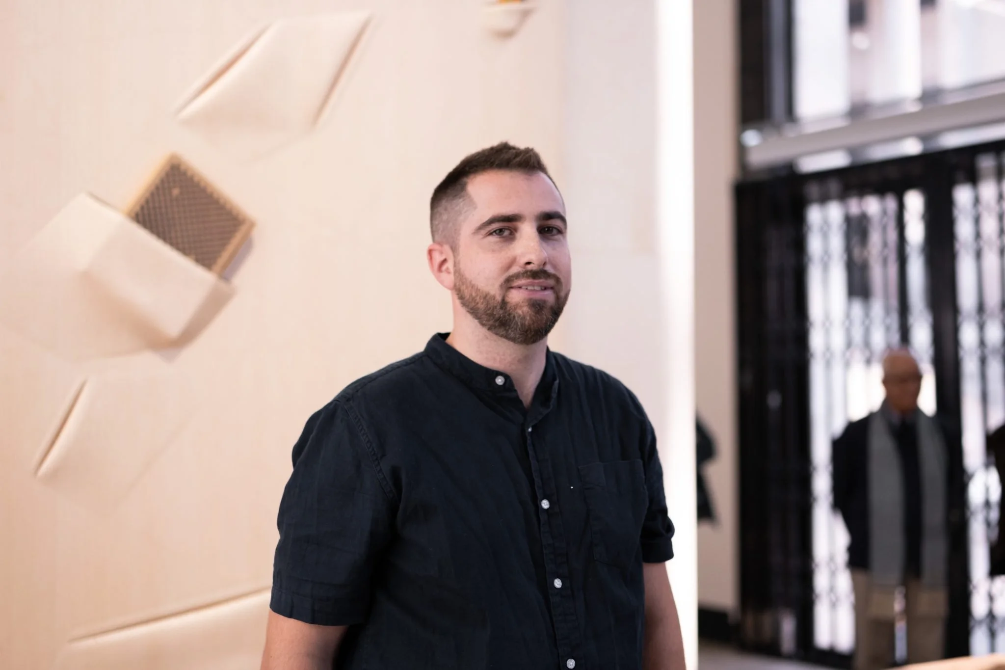 Un homme avec une barbe et une chemise sombre devant un mur beige avec des éléments décoratifs en relief. En arrière-plan, il y a un étage avec des personnes et une entrée avec des portes métalliques.