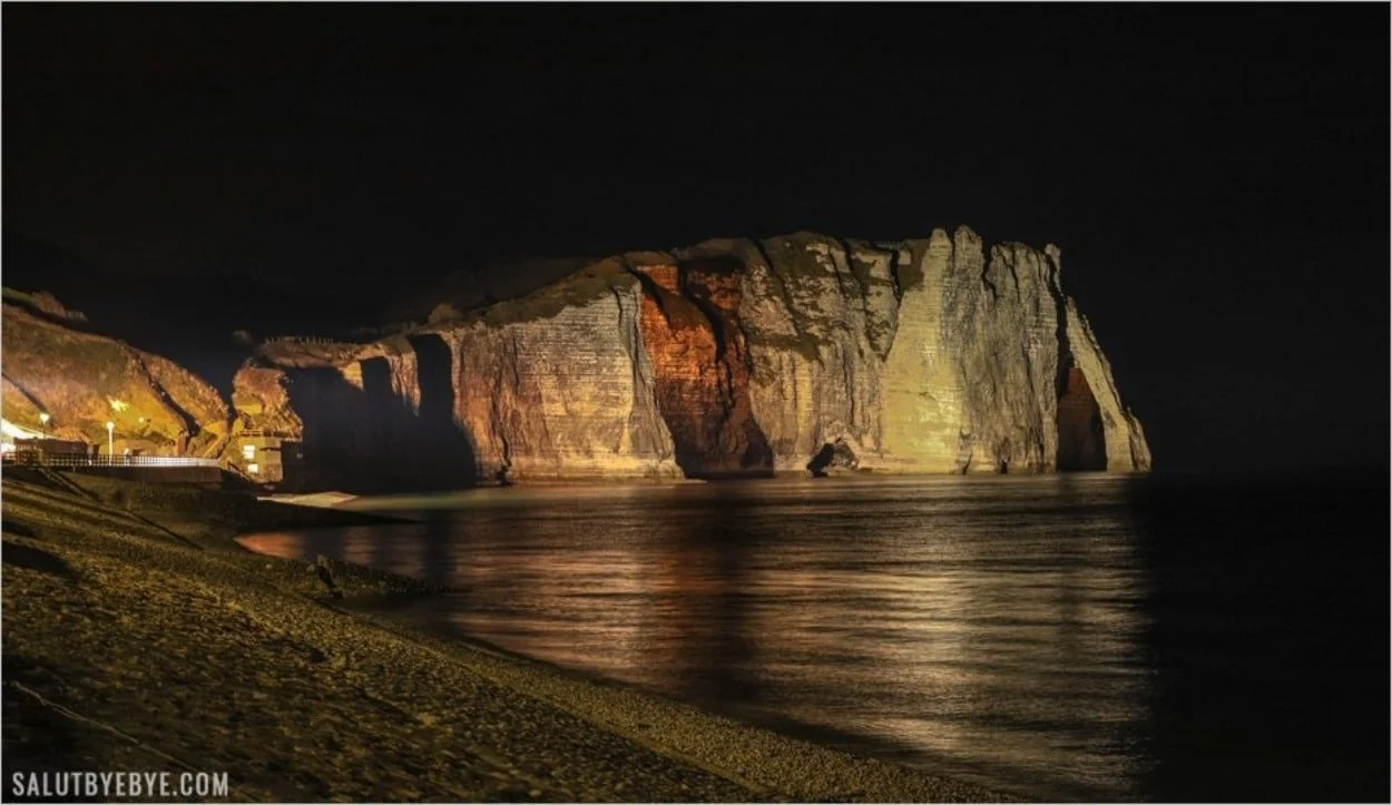 Côte rocheuse avec falaises illuminées la nuit, reflet dans la mer calme, plage sombre en premier plan, éclairage artificiel sur la gauche.