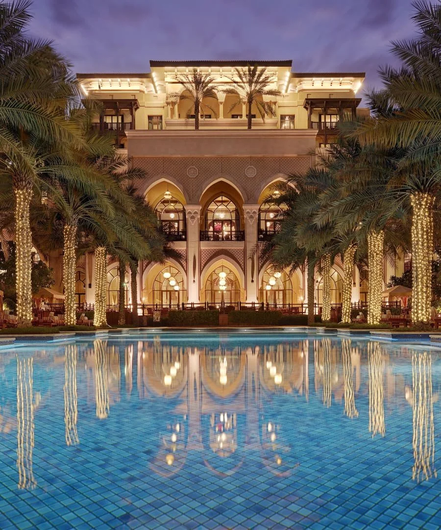 Un grand bâtiment éclairé avec des arcs islamique, entouré de palmiers décorés de lumières, avec une piscine devant reflétant la structure, en soirée ou la nuit.