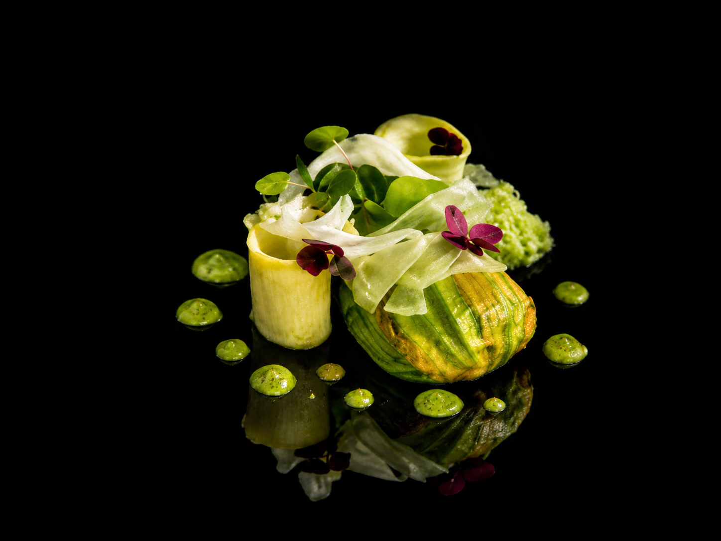 Présentation gastronomique d'un plat en salade avec des morceaux de courgettes, des feuilles vertes et violettes, et des petites gouttes vertes, sur fond noir.