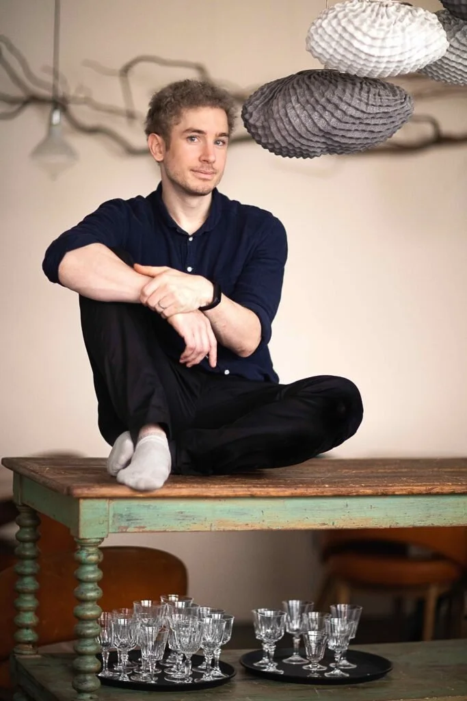 Un homme assis en tailleur sur une table en bois, portant un pantalon noir, une chemise bleue et des chaussettes blanches, dans une pièce avec des lampes en papier suspendues au plafond, et des verres à liqueur sur une table en dessous.
