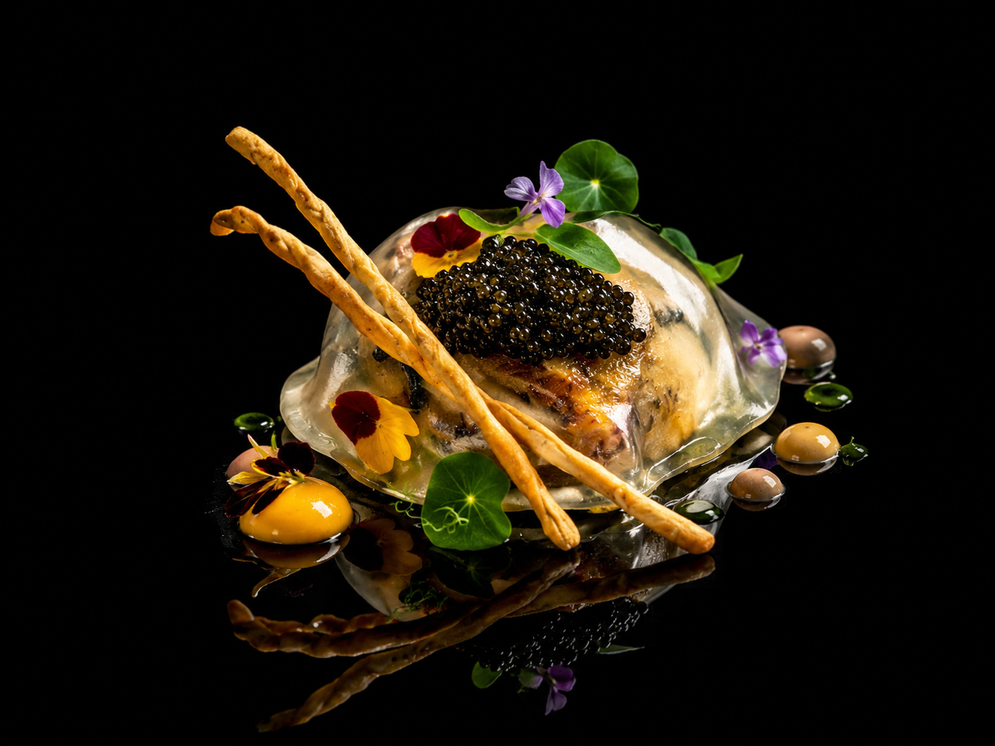 Plat gastronomique avec gelée transparente, caviar noir, fleurs violettes et jaunes, deux baguettes croustillantes, et diverses petites décorations végétales, sur fond noir.