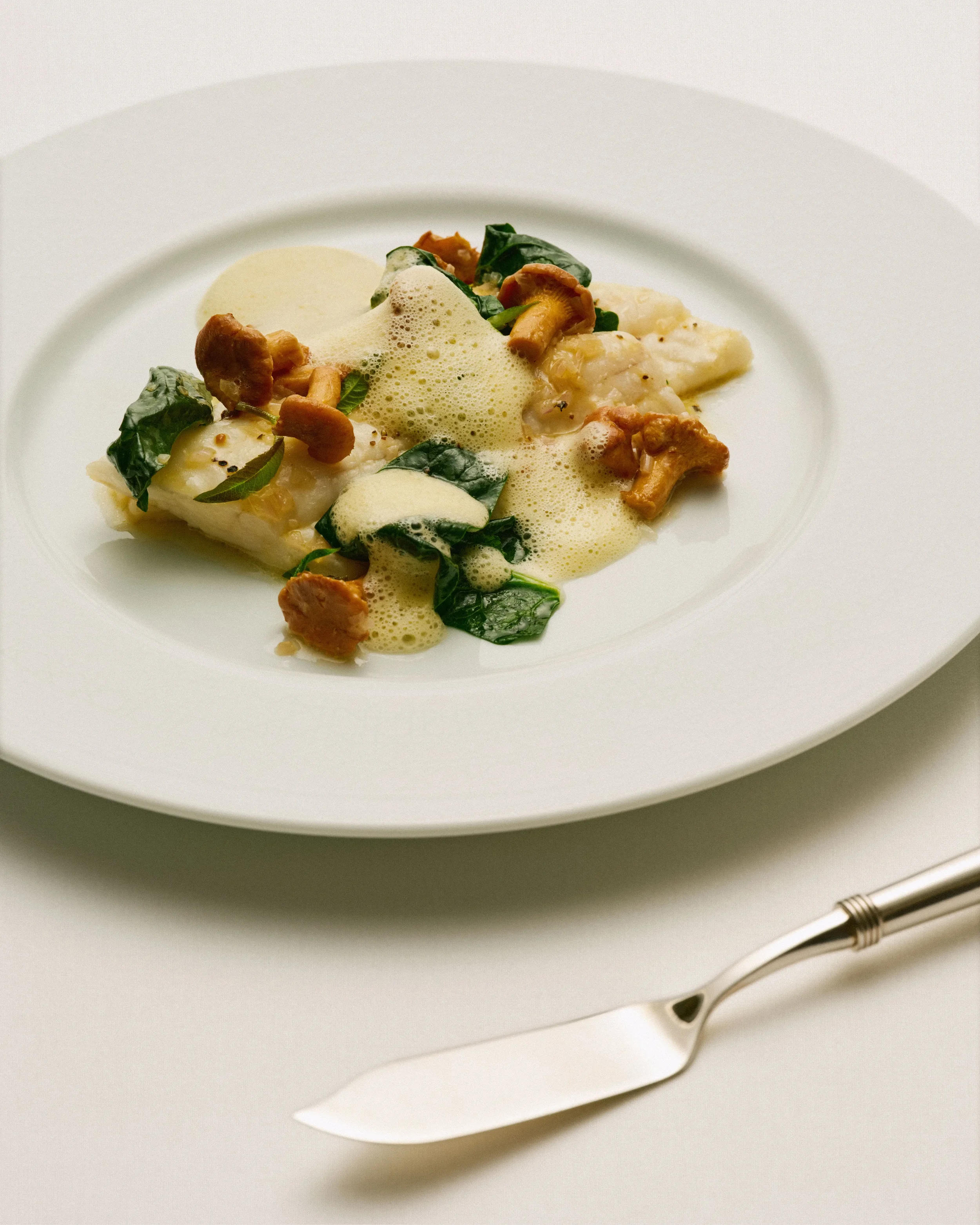 Plat gastronomique avec purée, champignons, épinards et mousse, présenté sur une assiette blanche avec une cuillère à dessert argentée à côté.