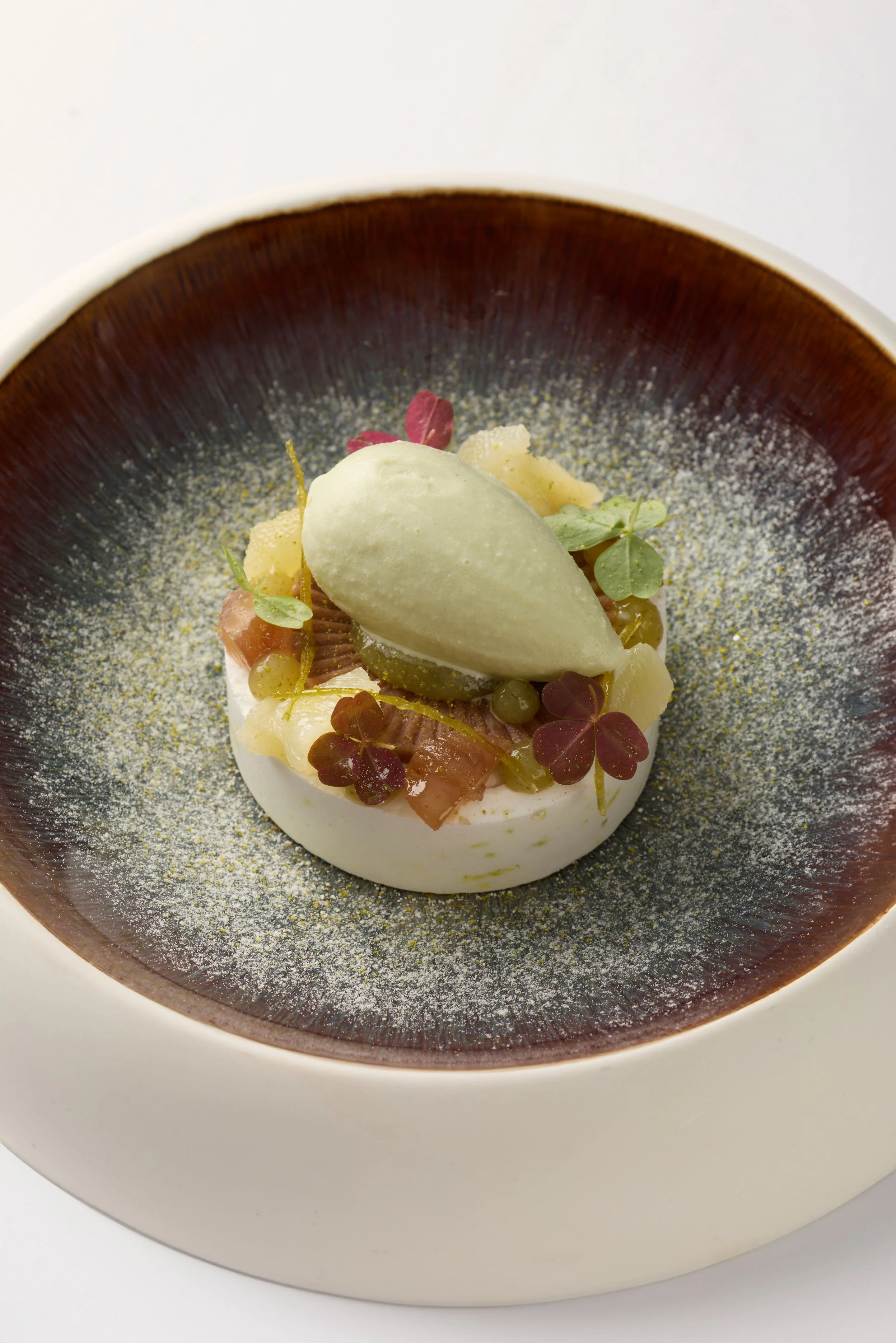Dessert sophistiqué avec une quenelle de glace verte sur un lit de fruits, de petits morceaux de gâteau ou biscuit, décoré de microgreens, servi dans un bol en céramique.