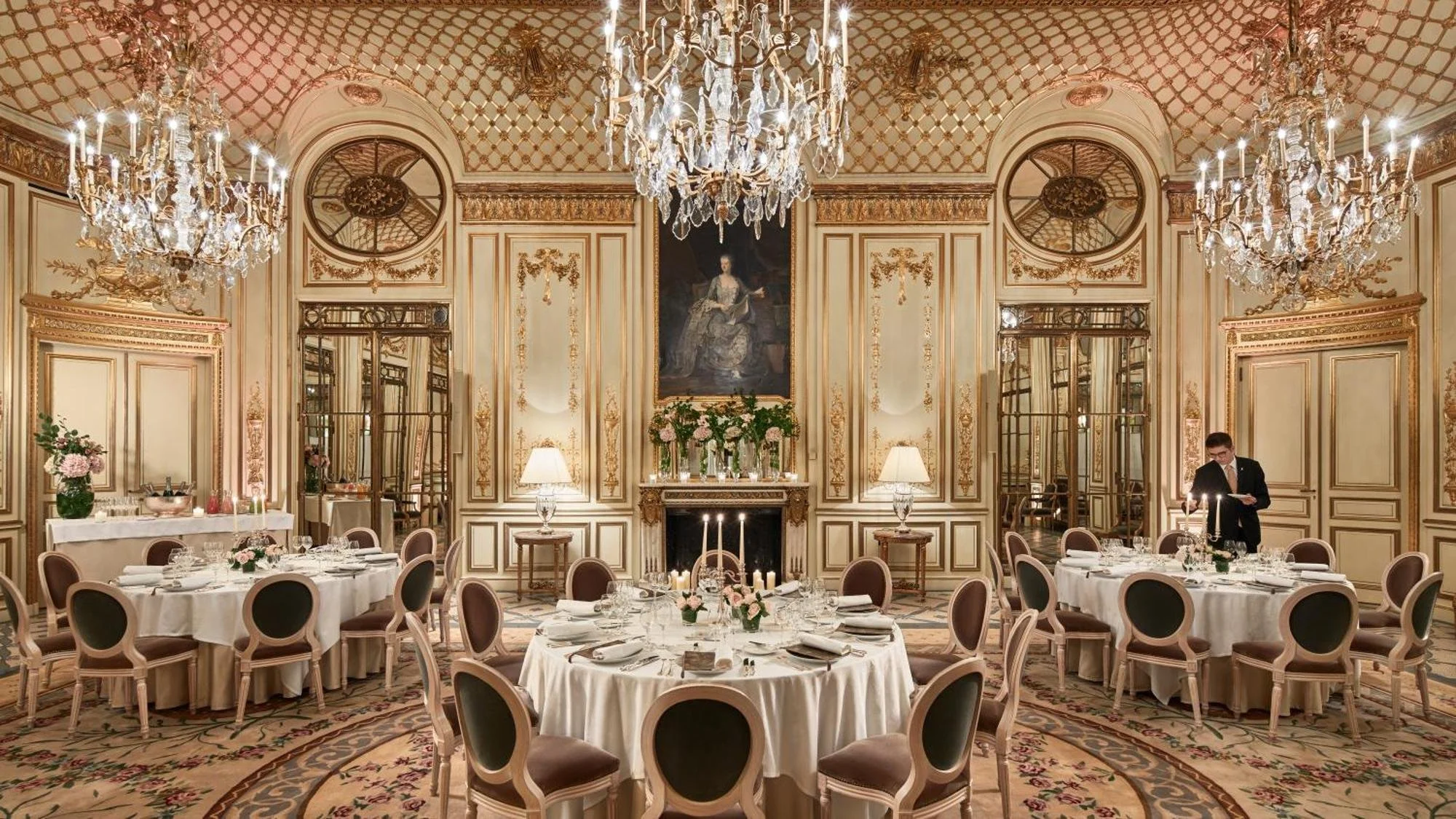 Salle à manger élégante avec chandeliers, murs décorés en style classique, tables rondes avec linge blanc, vaisselle et fleurs, un serveur en train de préparer une table.
