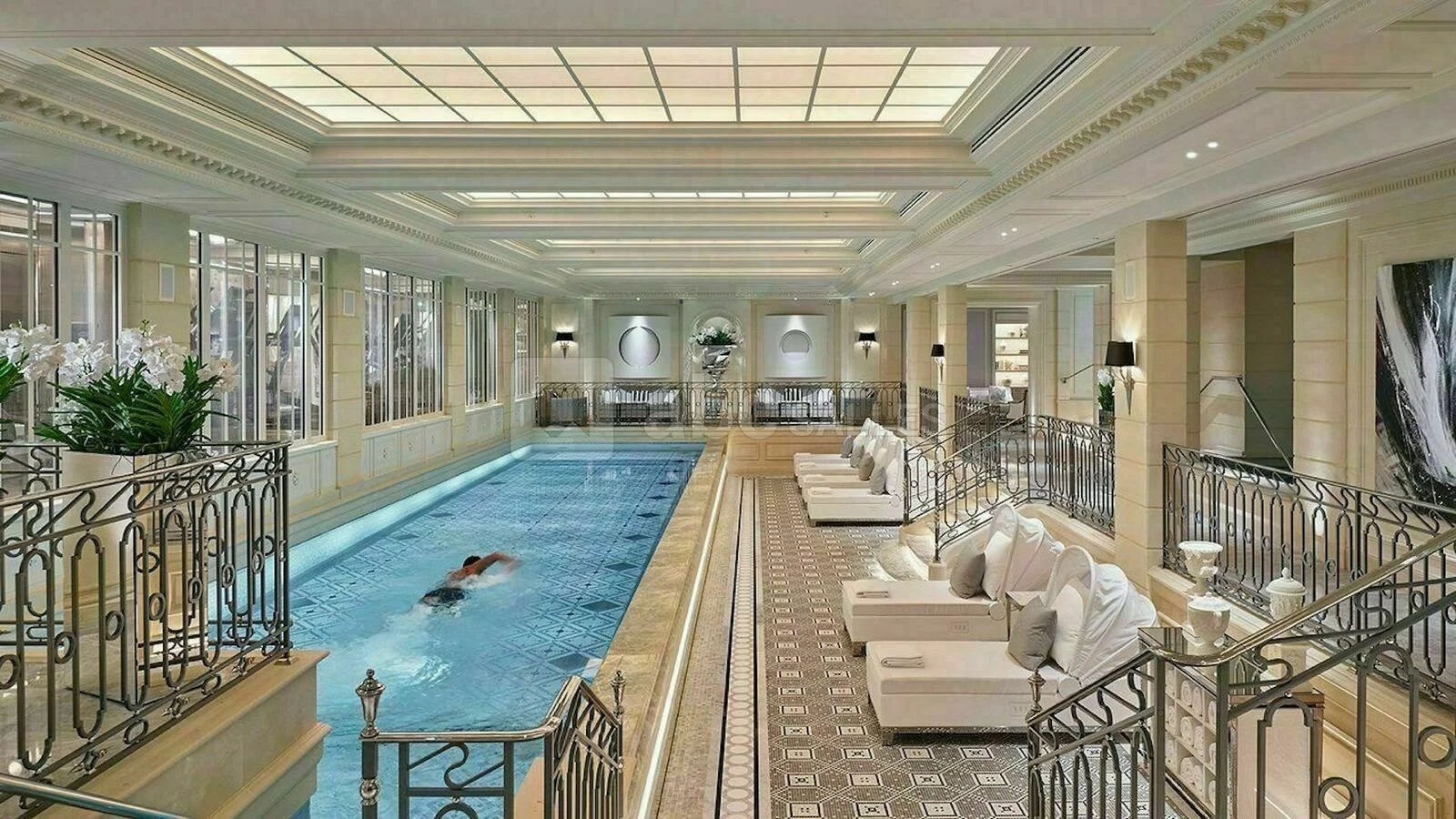 Piscine intérieure luxueuse avec un nageur, fauteuils blancs avec des coussins, décor élégant, grandes fenêtres, plantes et luminaires suspendus.