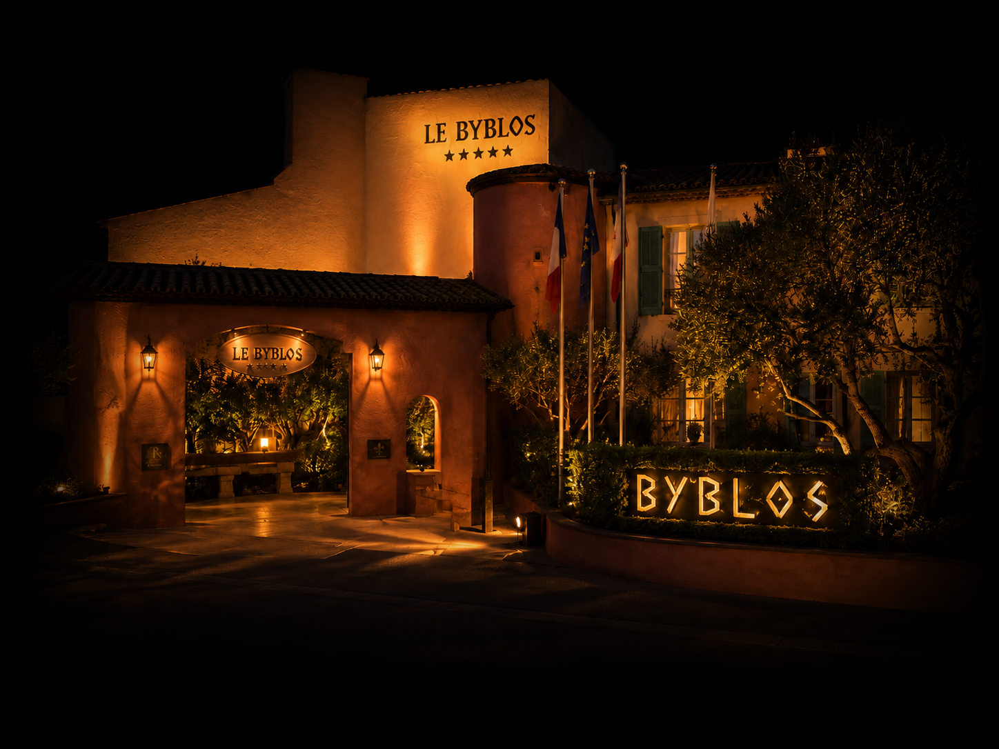 Une façade d'hôtel appelée 'Le Byblos' éclairée de nuit, avec une entrée ornée de lumières, un panneau lumineux indiquant 'Byblos', et trois drapeaux (français, européen et un autre), entourée de végétation et éclairée par des lampes.