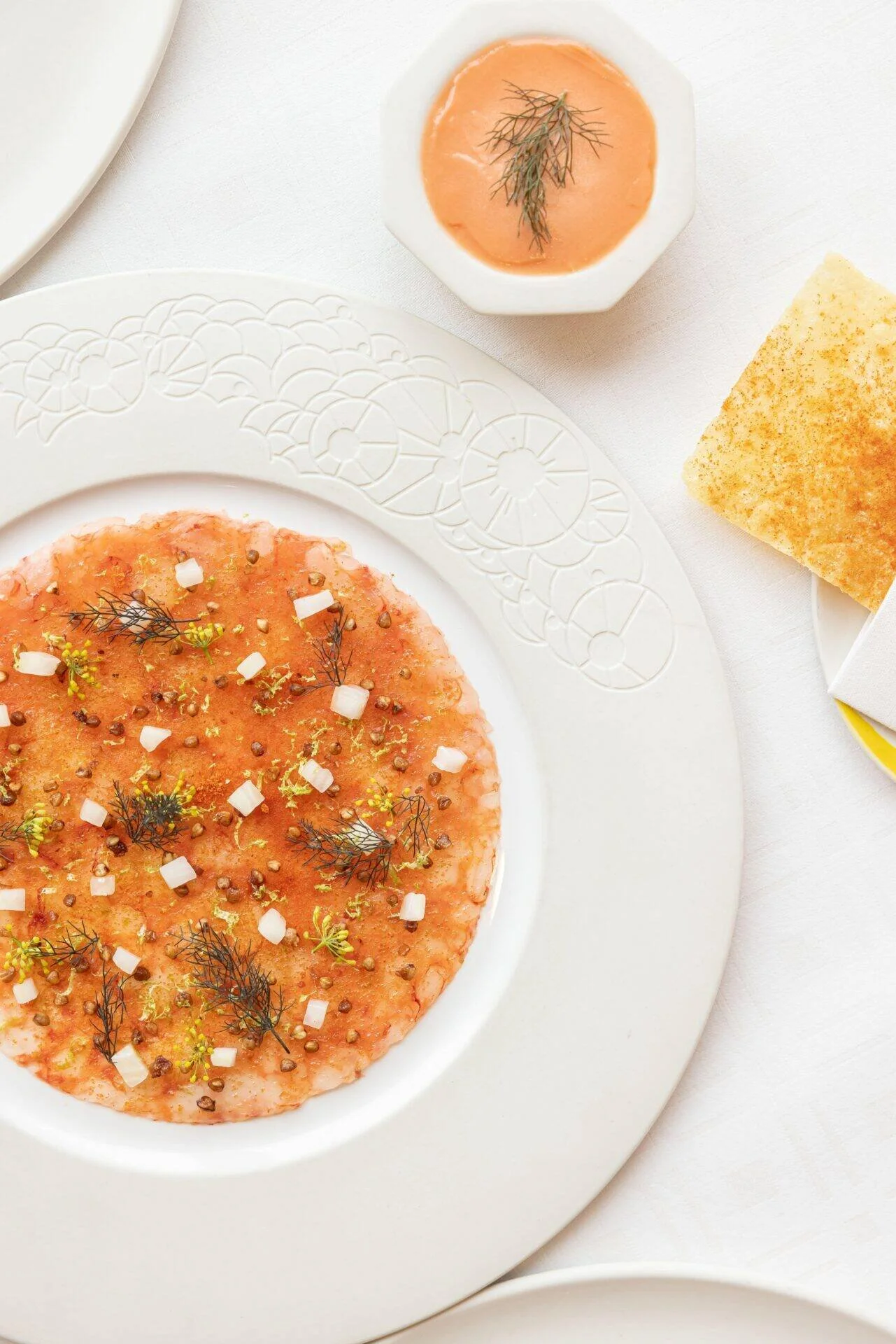 Une assiette de carpaccio de saumon garni de fines herbes, avec une sauce rose en petit bol, un morceau de pain croustillant et un carré de beurre sur une table blanche.