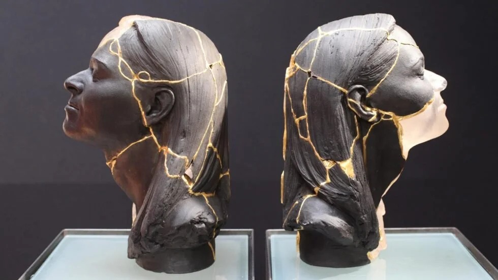 Deux bustes de femmes en plastique noir endommagés avec des fissures dorées, exposés sur des supports en vitrine.