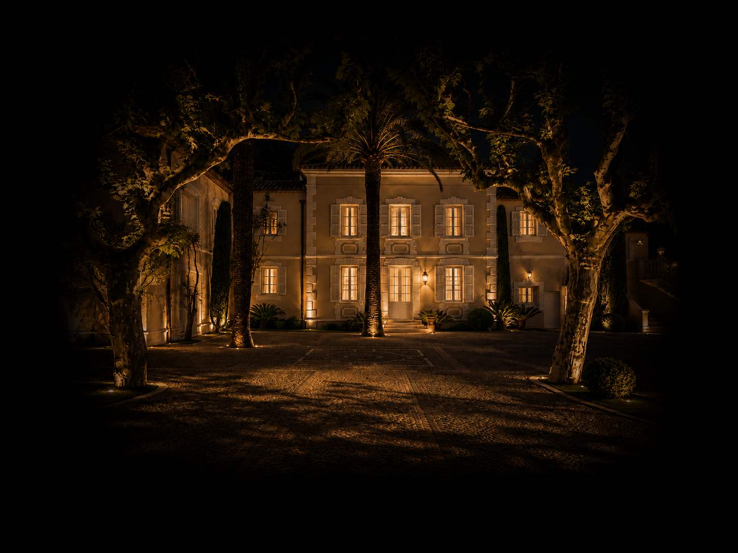 Une grande maison éclairée la nuit, entourée d'arbres avec des lumières au sol, en façade