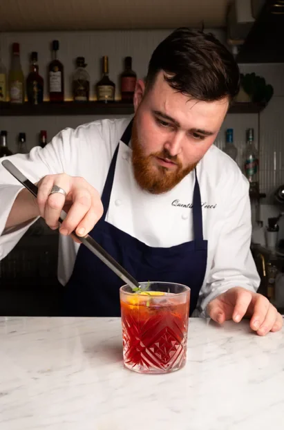 Un barman prépare un cocktail rouge dans un verre en cristal, en utilisant une cuillère de décoration, derrière lui des bouteilles d'alcool sur une étagère.