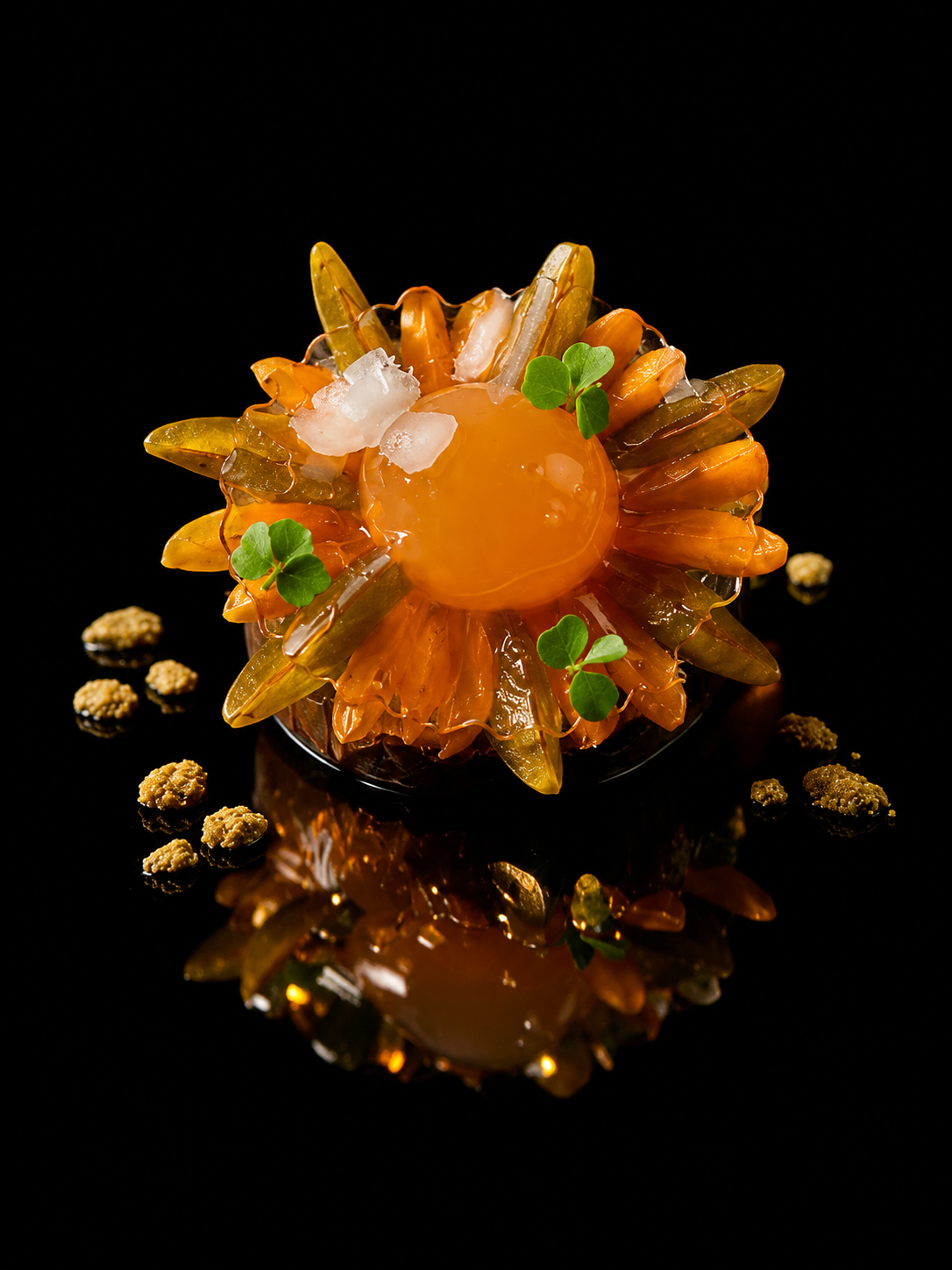 Dessert artistique avec une sphère orange au centre, entourée de tranches translucides d'orange et décorée de microgreens, sur un fond noir avec une réflexion