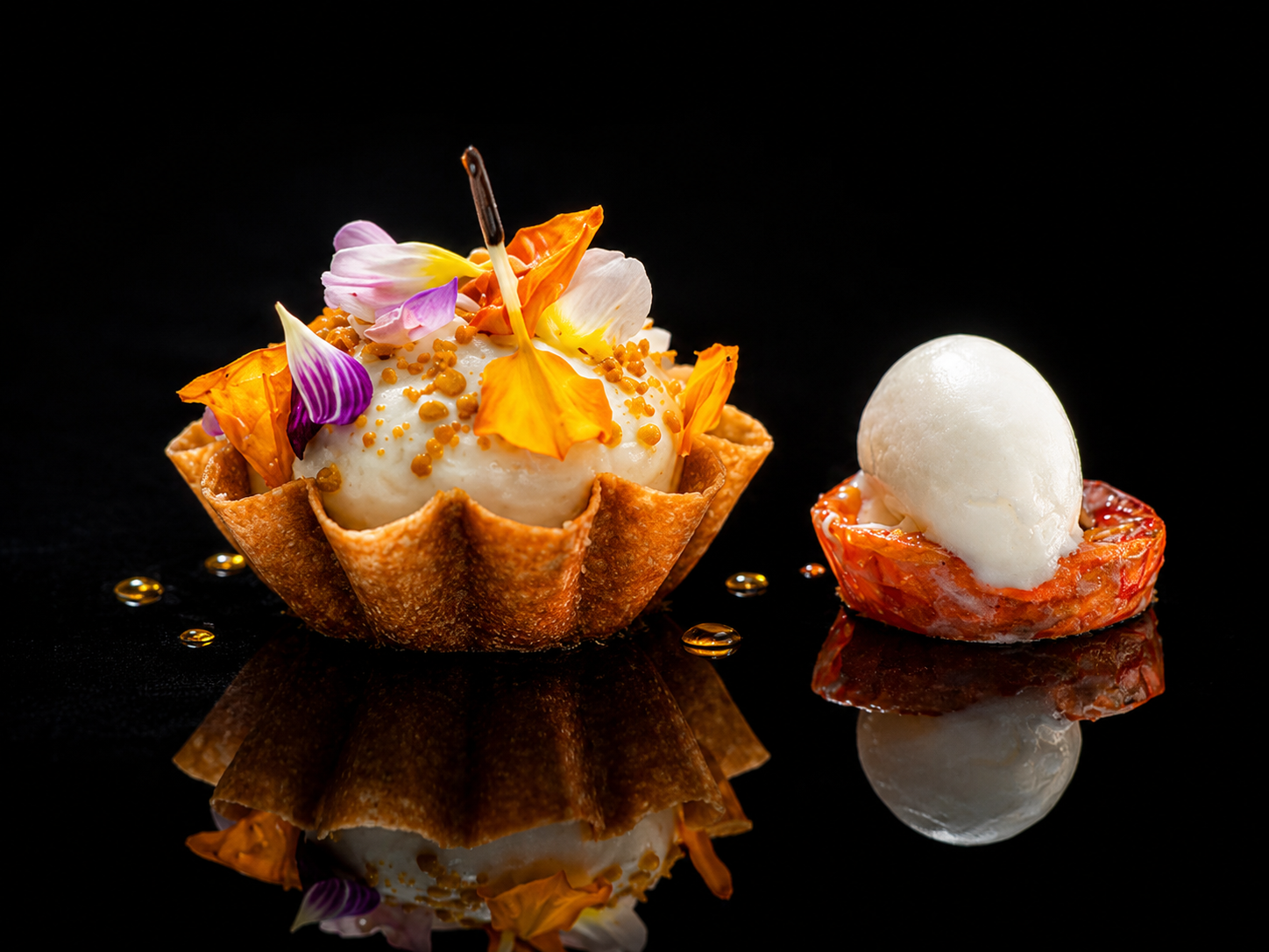 Deux desserts élégants avec glace à la vanille et des fleurs comestibles, un dans une coupe en cornet de biscuit, et l'autre dans un petit bol, sur un fond noir brillant.
