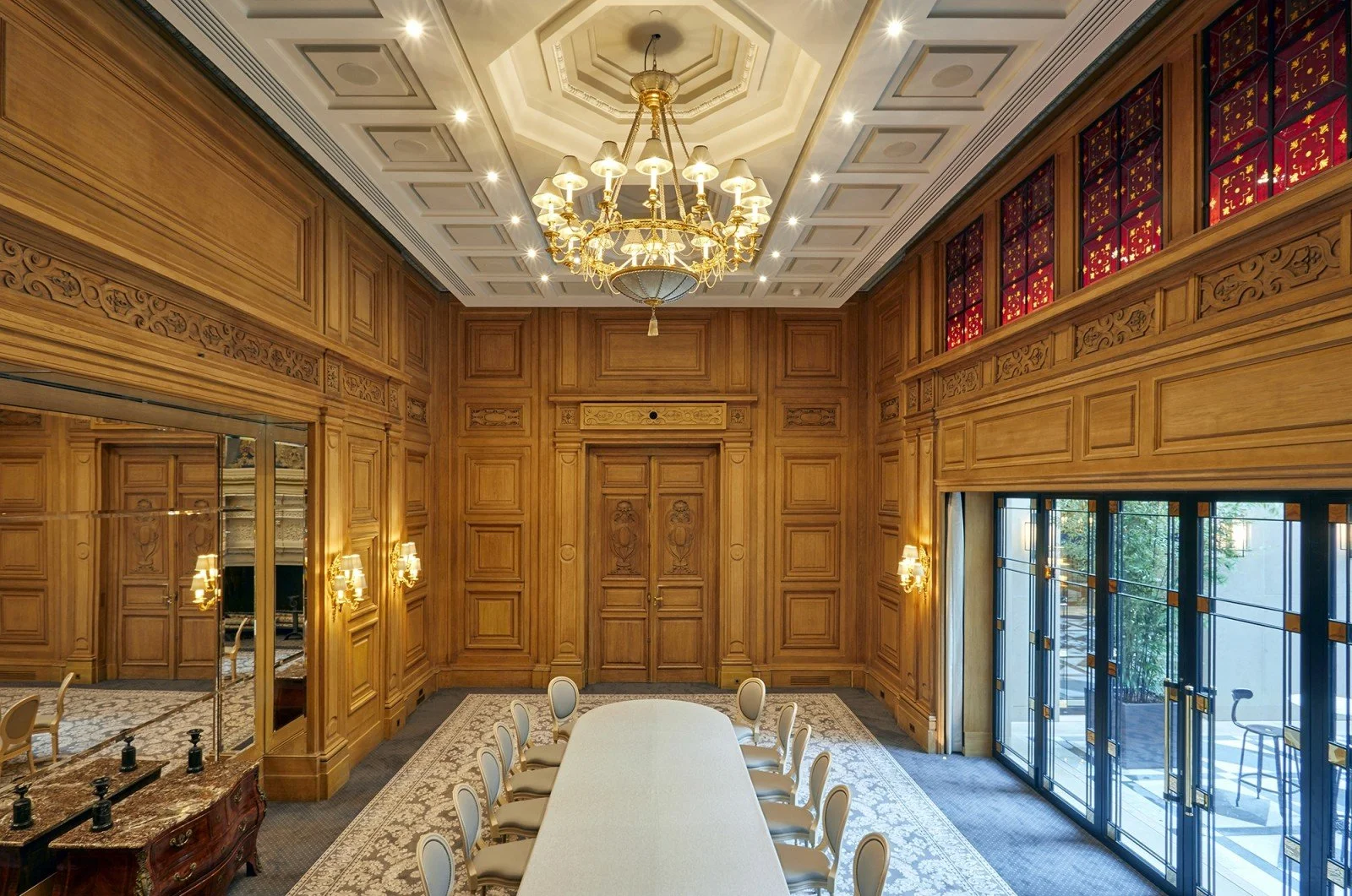 Une salle de réunion élégante avec des murs en bois sculpté, une longue table blanche entourée de chaises, un chandelier doré suspendu au plafond, et des fenêtres en verre donnant sur un patio.