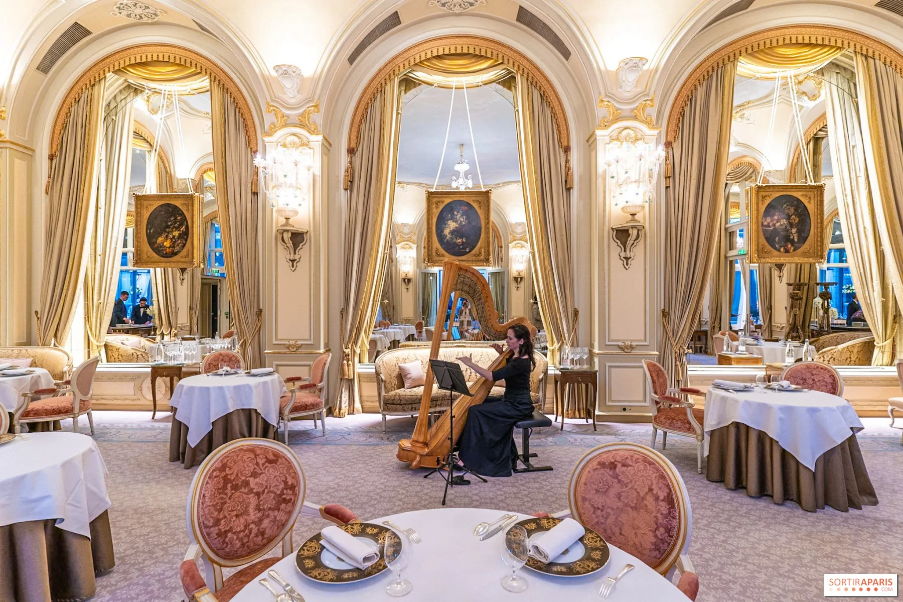 Une salle de restaurant élégante avec une harpiste jouant au centre. La pièce est décorée de rideaux dorés, de chandeliers et de peintures florales. Les tables sont dressées avec des nappes blanches, des assiettes noires et des couverts en argent.