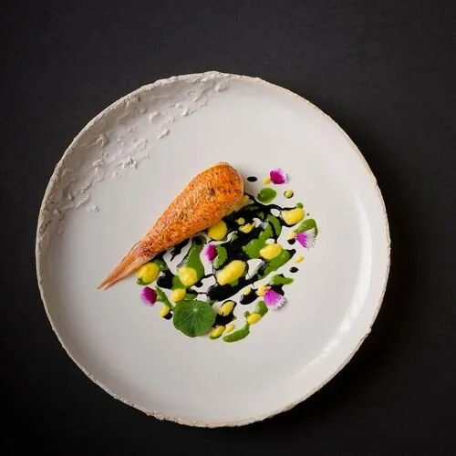 Assiette de présentation avec un filet de poisson croustillant, accompagnée de sauces colorées, de feuilles vertes, et de petites fleurs violettes.