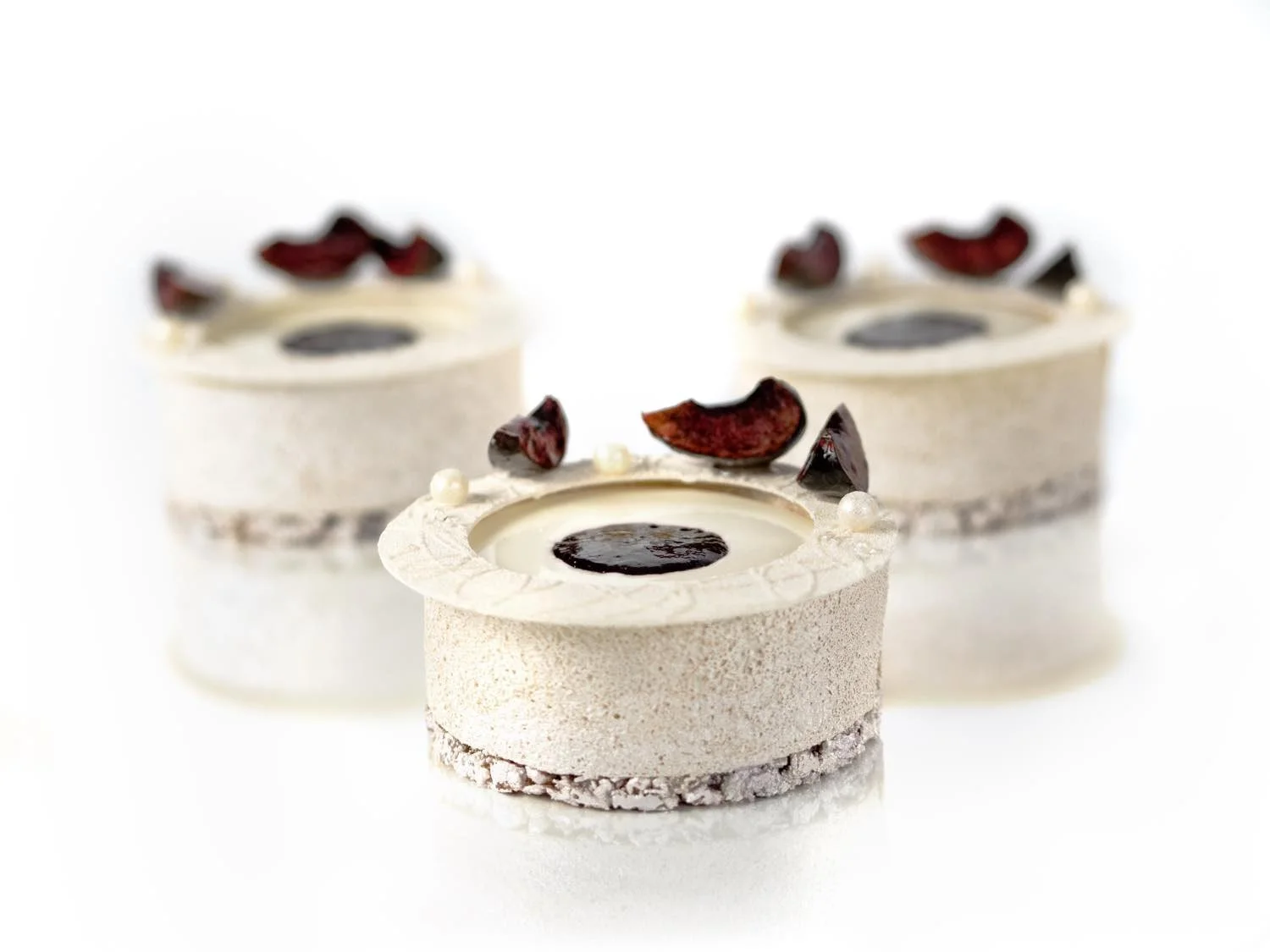 Trois petits gâteaux blancs ronds décorés de morceaux de fruit rouge et de perles blanches, avec un centre de chocolat liquide, sur un fond blanc.