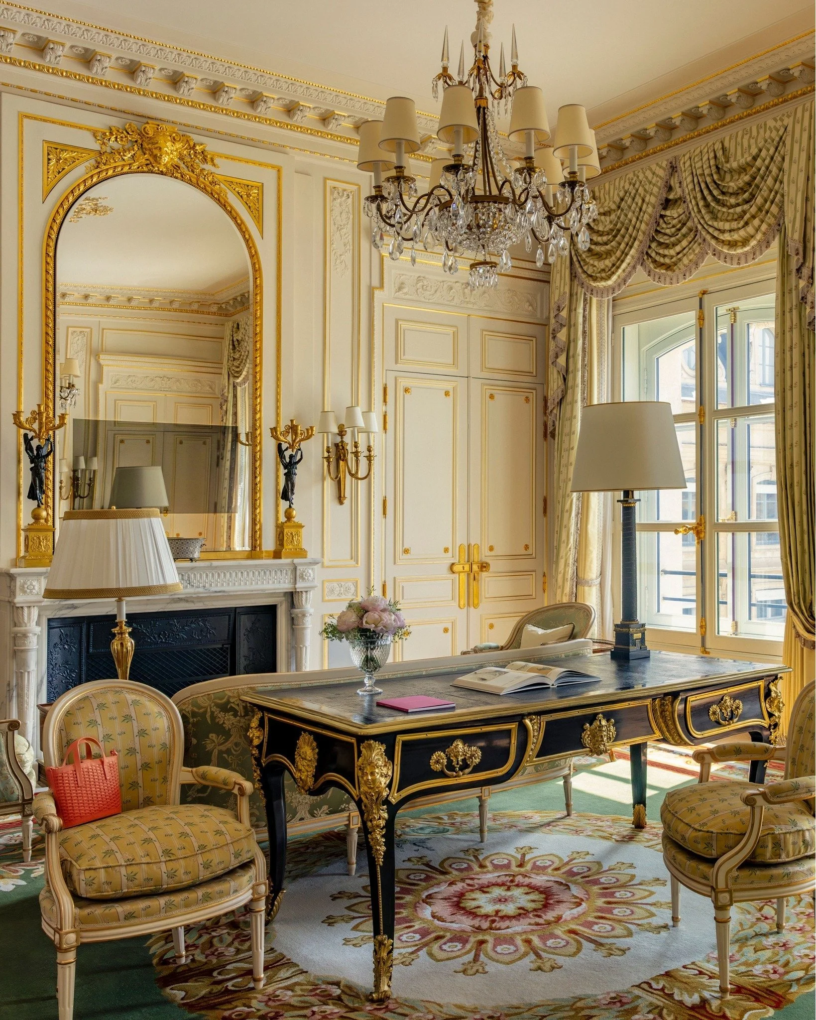Salon élégamment décoré avec un grand chandelier, un miroir ancien avec cadre doré, une cheminée blanche, un bureau noir avec dorures, une lampe de bureau, fauteuils en bois, rideaux à motifs et un tapis floral.