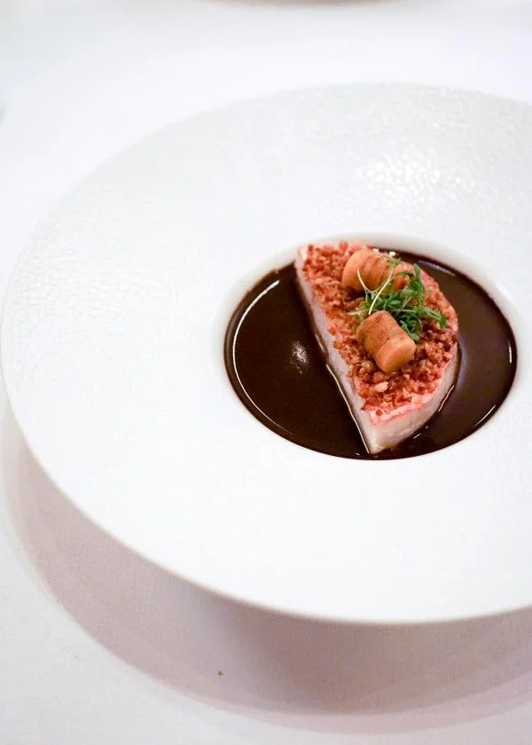 Dessert présenté dans un bol blanc avec une sauce chocolatée, garnie de morceaux de biscuit, de micro-pousses et de rouleaux de chocolat.