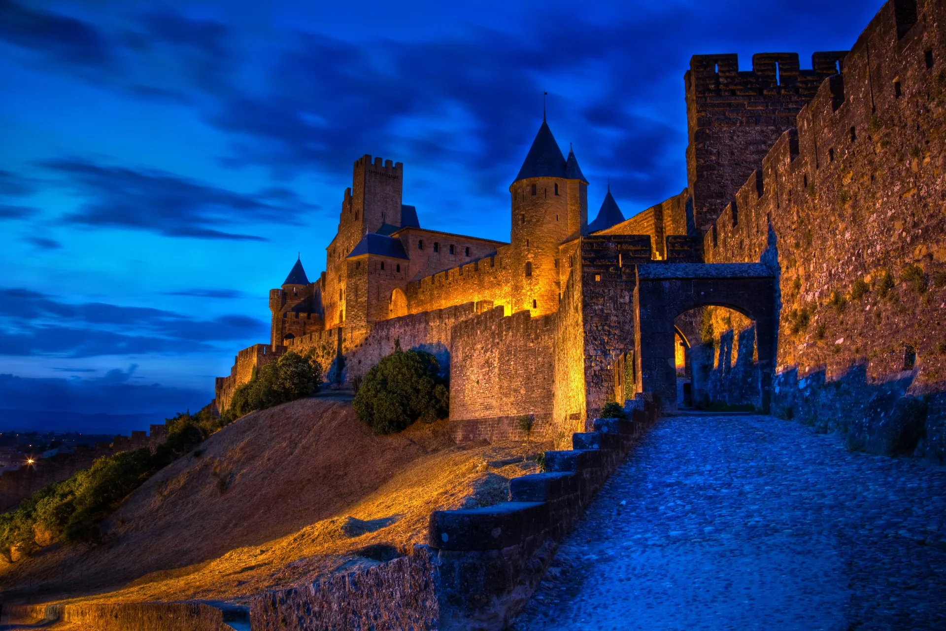 Château médiéval illuminé la nuit avec un ciel bleu et nuages, situé sur une colline, entouré de murs et de toits pointus, avec une voie pavée en premier plan.