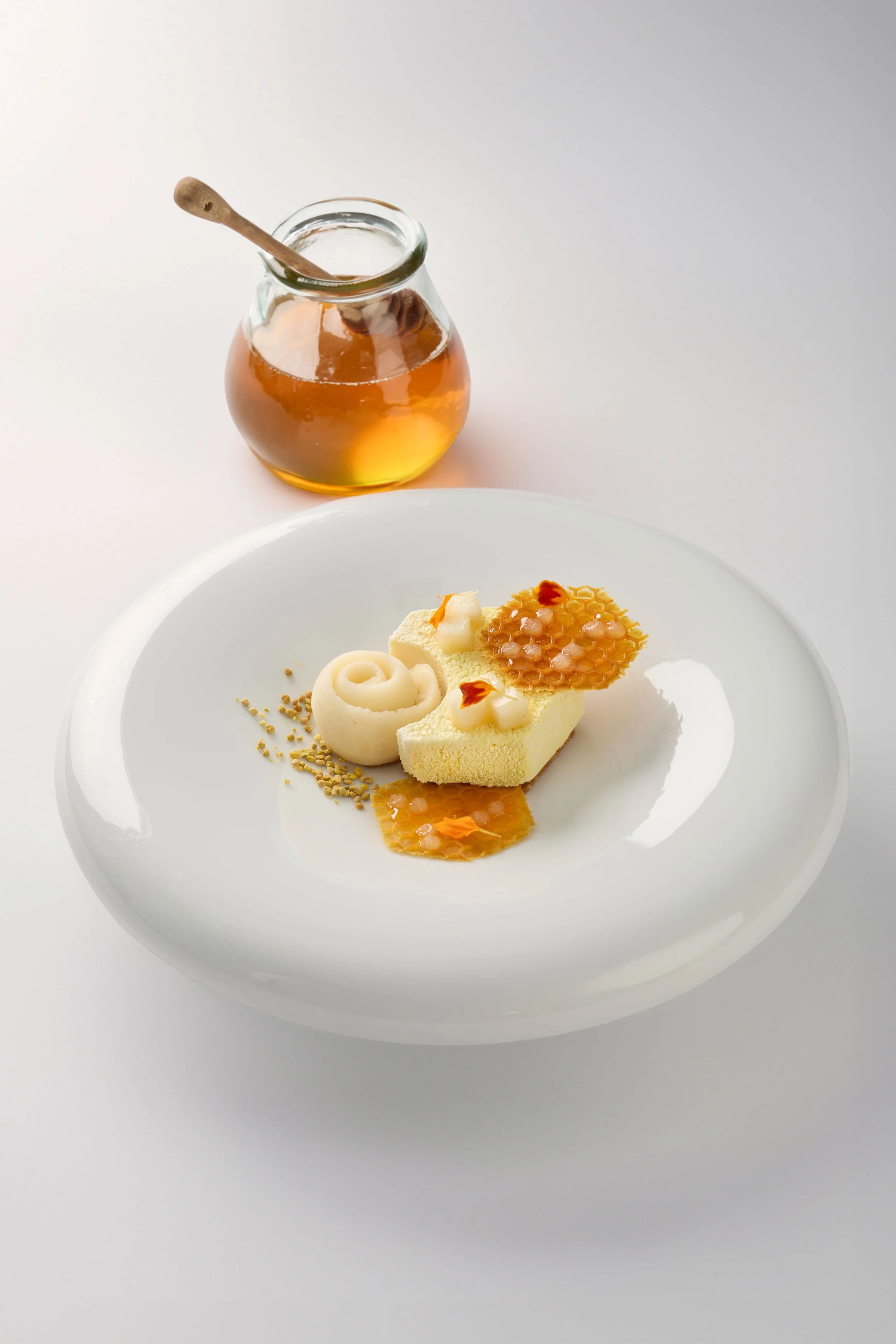 Dessert sophistiqué avec un support de crème, un décor en miel et un morceau de miel sur une assiette blanche, accompagnée d'un pot de miel avec une cuillère en bois.