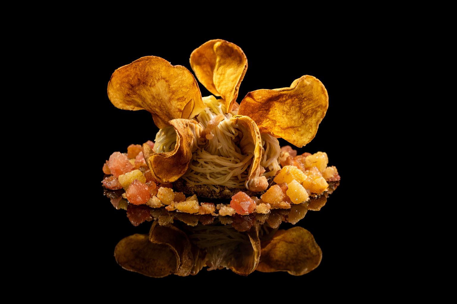 Un plat sophistiqué avec des nouilles nues au centre, entourées de morceaux de fruits ou légumes colorés, avec une garniture de chips de pommes de terre ou de légumes déployés de façon artistique, présenté sur un fond noir.