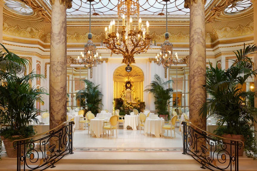 Une salle de réception élégante avec des tables rondes, des chaises jaunes, de grands lustres en cristal, des colonnes en marbre, et des plantes vertes décoratives, avec un décor doré et classique.