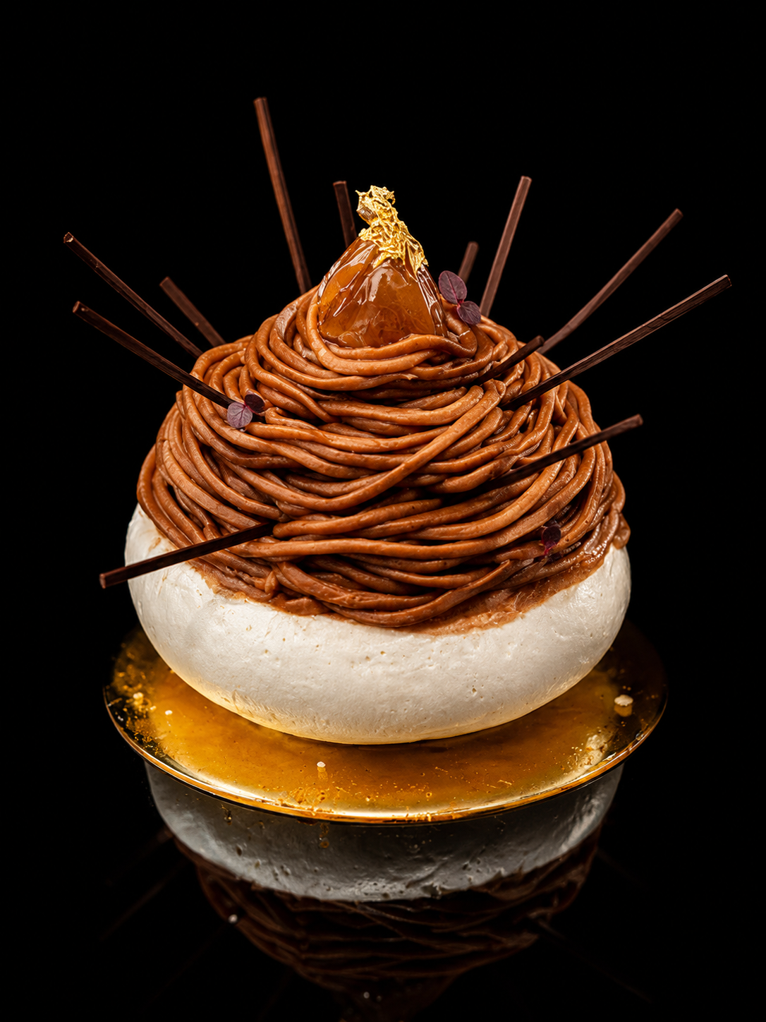 Un dessert sophistiqué avec une base blanche ronde, garnie de twiste de chocolat ressemblant à des spaghettis, décoré avec une pièce de caramel dorée, une sphère de caramel ou fruit, et des fines tiges de chocolat ou biscuit, présenté sur une plaque dorée sur fond noir.