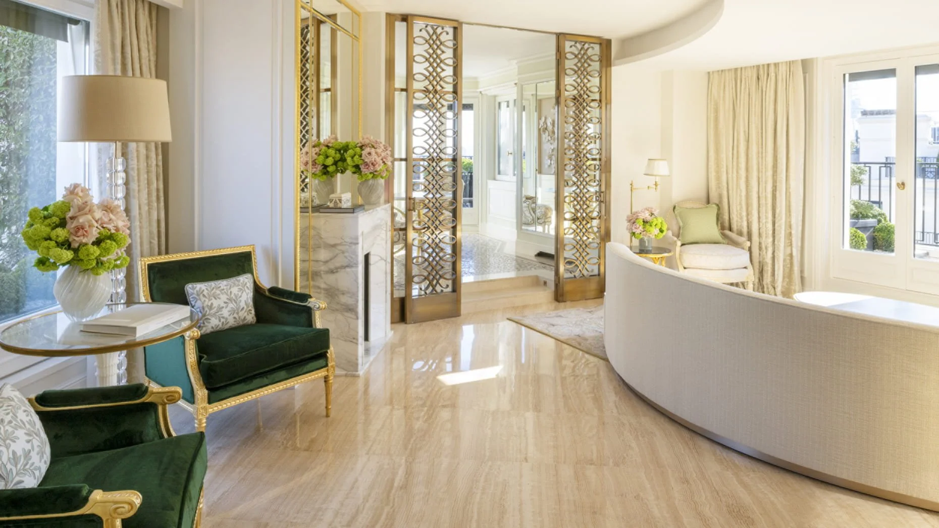 Salon lumineux avec fauteuils en velours vert, vases de fleurs, grandes fenêtres avec rideaux beige, miroir, porte en treillage doré séparant la pièce
