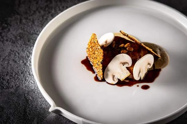 Assiette blanche avec un dessert composé de sauce chocolat, tranches de champignons, une tuile croustillante et des graines
