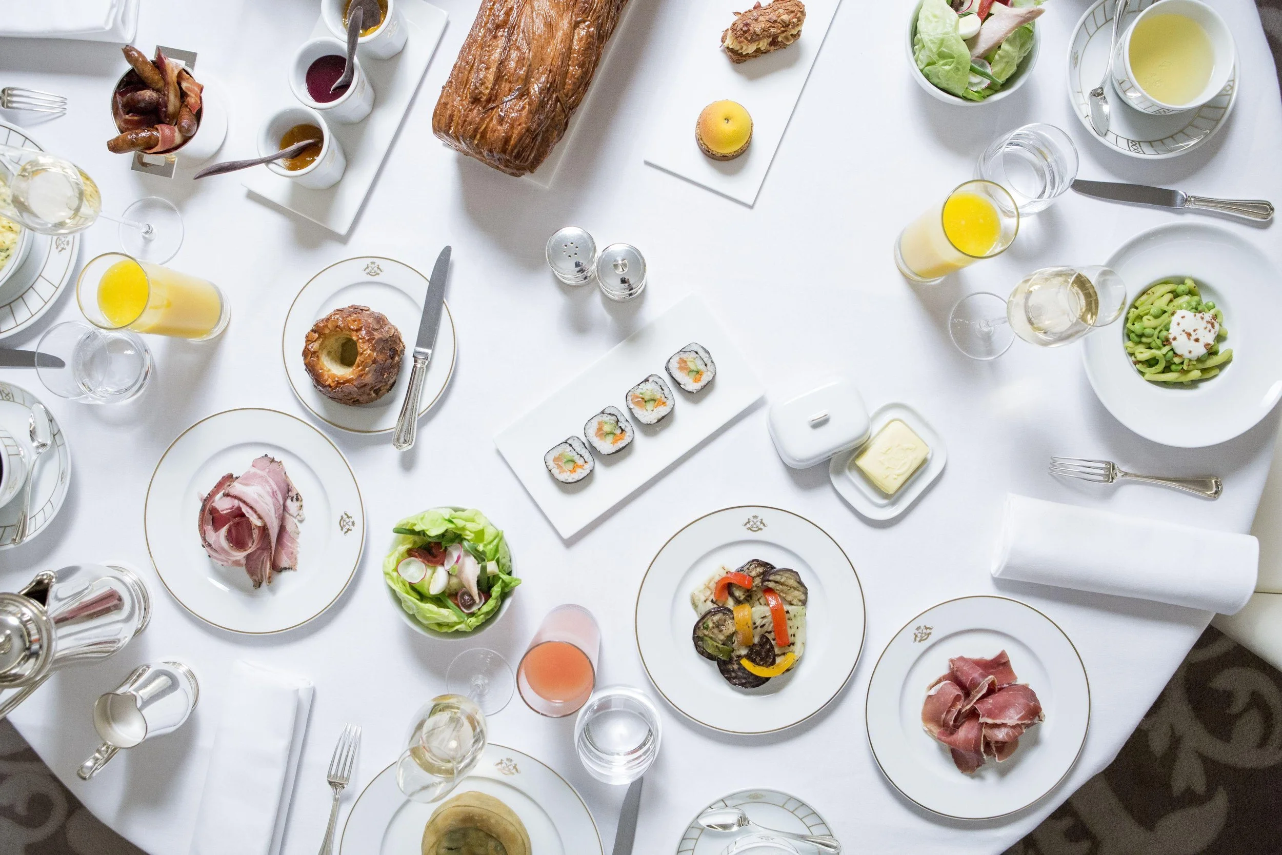 Table dressée pour un repas avec divers plats, boissons et desserts, notamment des sushis, du pain, des salades, du foie gras, du jambon, des smoothies, du vin blanc, et différentes sauces.