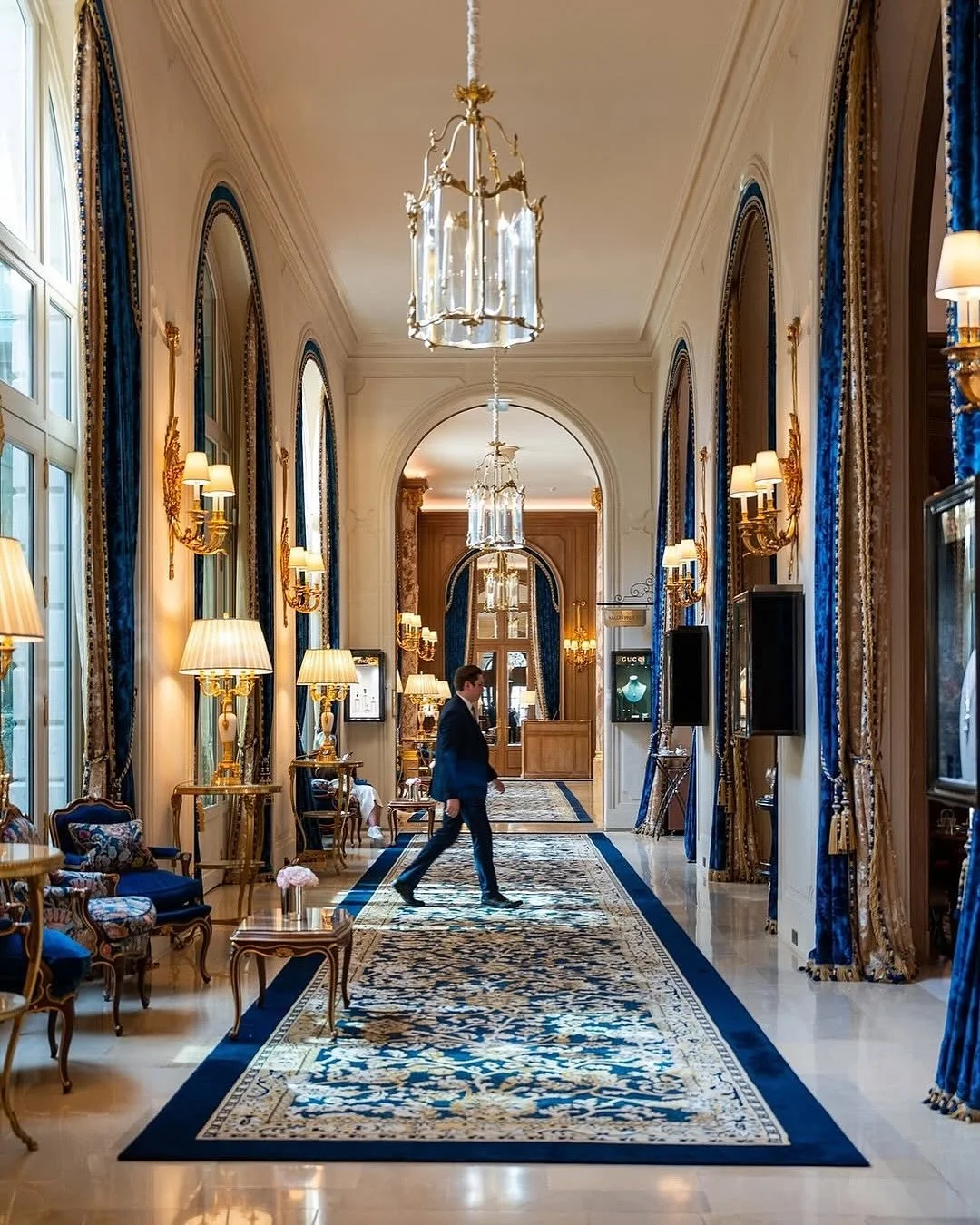 Un long corridor luxueux avec des murs ornés de moulures, des fenêtres avec des rideaux bleus et dorés, des lustres en cristal suspendus au plafond, un tapis aux motifs floraux, et des lampes à abat-jour. Un homme en costume marche au centre.