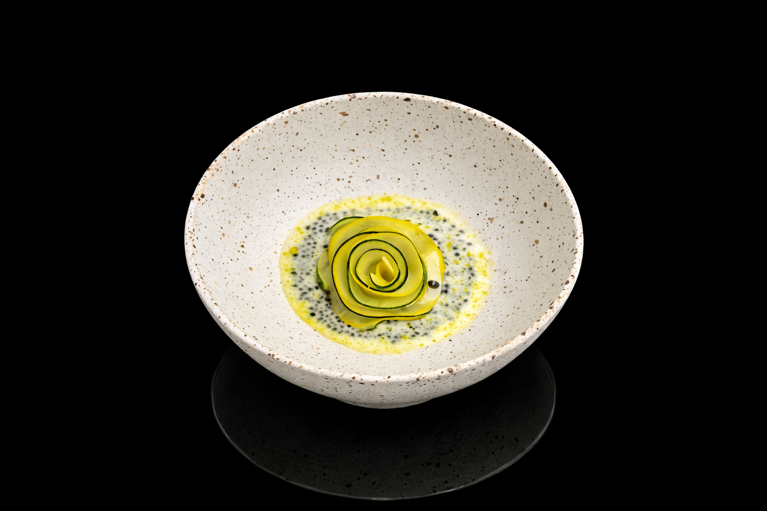 Assiette avec un rouleau de courgettes en spirale napée d'une sauce crémeuse aux grains noirs, sur un fond noir.