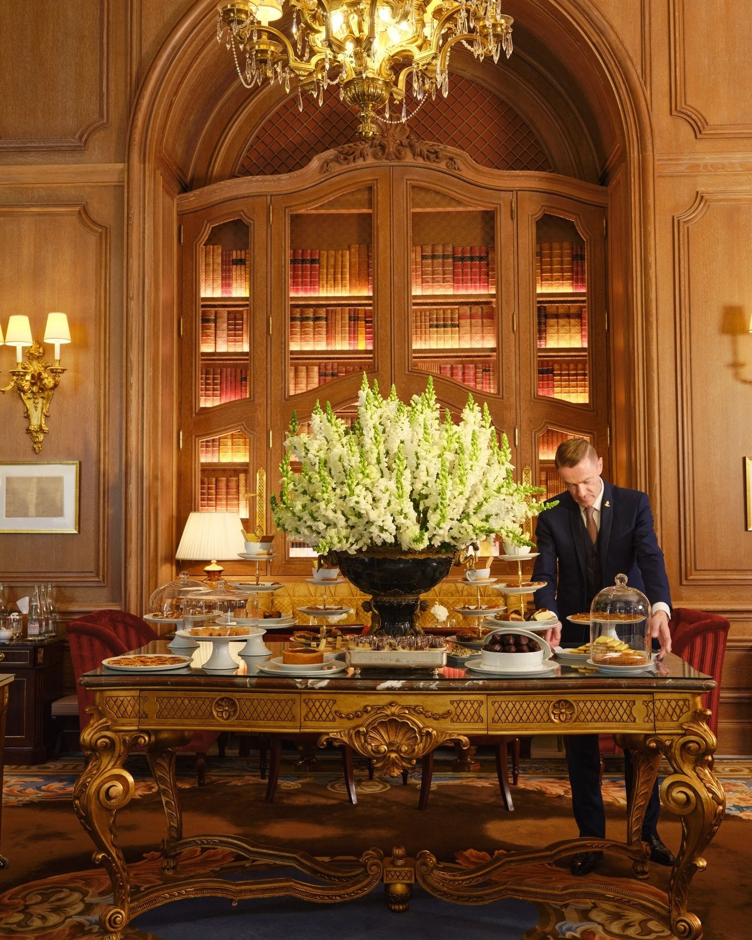 Salle de réception élégante avec un mobilier en bois, un plafonnier en cristal et une grande table ornée de fleurs blanches et de desserts.
