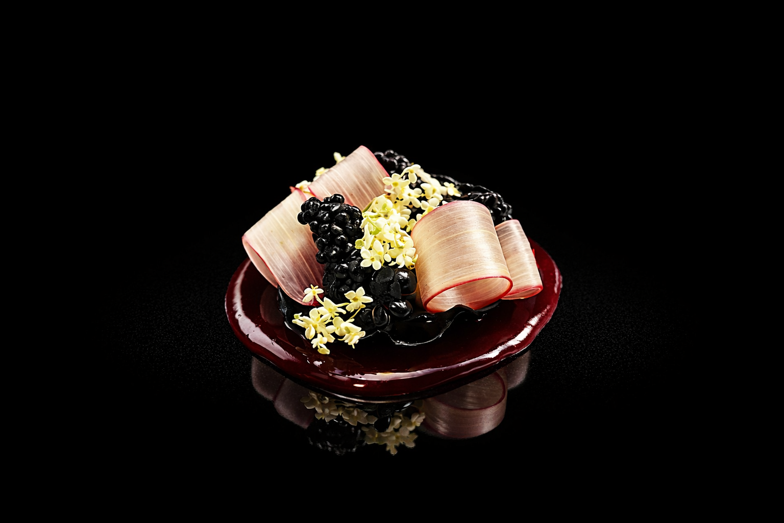Un plat de dessert sophistiqué avec des roses de pâte, des fleurs comestibles, et des baies noires sur une assiette rouge, fond noir.