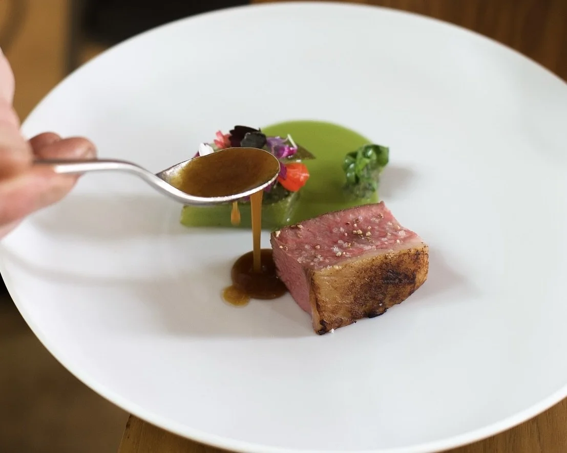Un filet de sauce brune est versé sur un morceau de viande saignant, accompagné de purée verte et de decoratifs légers