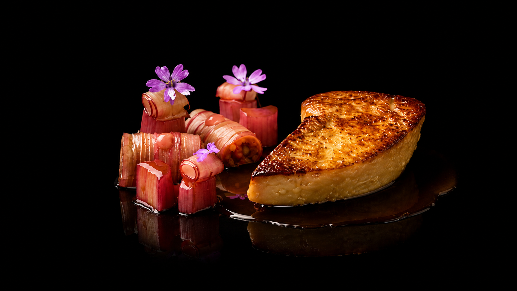 Une assiette avec un morceau de poisson grillé et des roulés de légumes garnis de petites fleurs violettes, présenté sur un fond noir élégant.