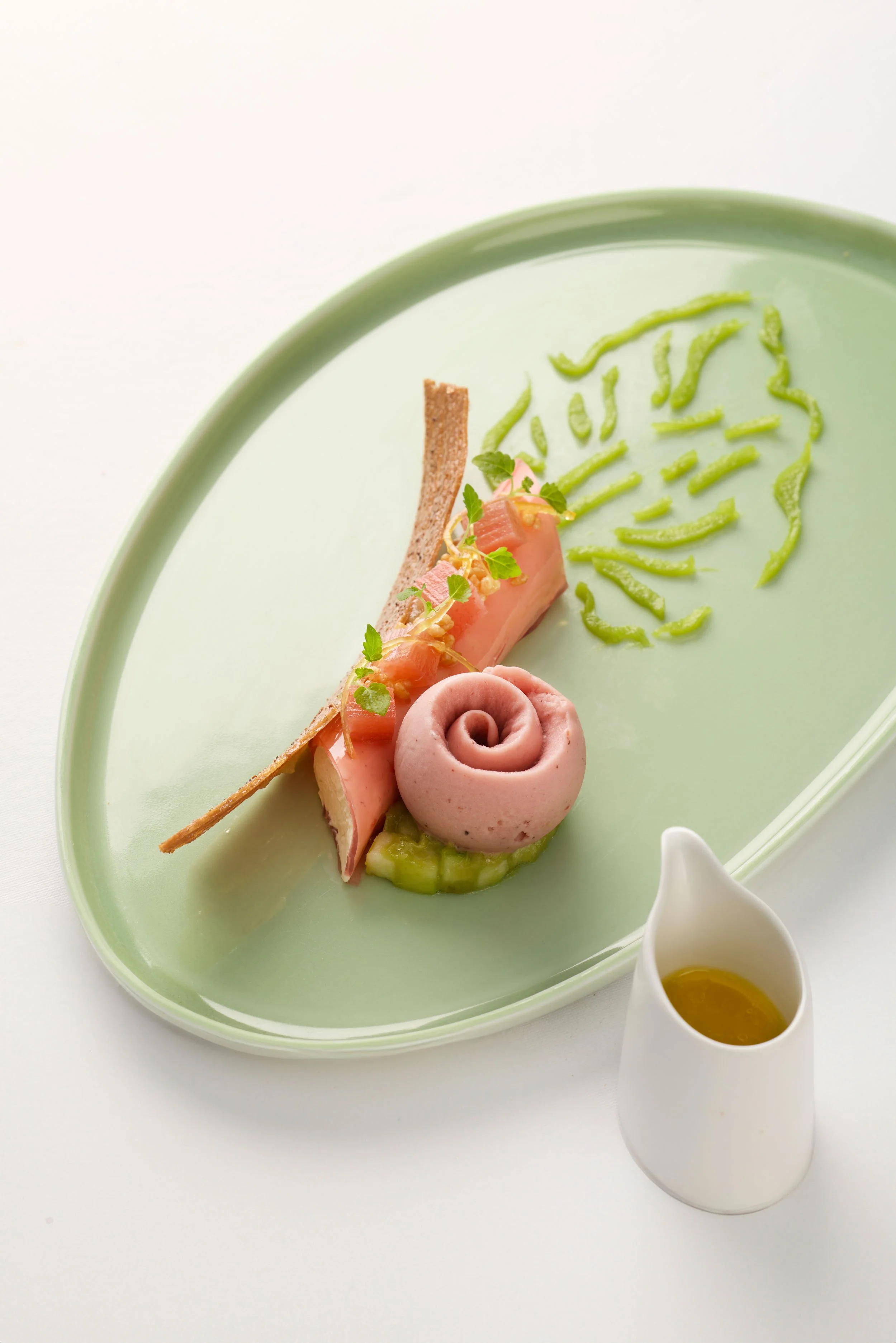 Une assiette de présentation avec du glaçage vert, un rouleau de charcuterie, une tranche de pain, une petite quantité de légumes et une sauce dans une petite cruche blanche.