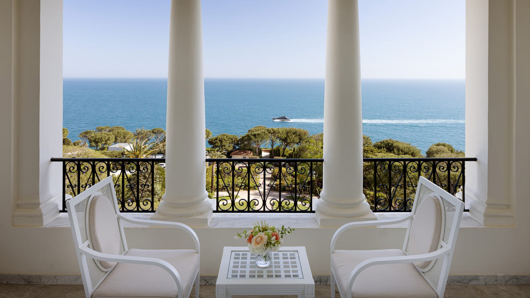 Vue d'une terrasse avec deux fauteuils blancs, une petite table avec un vase de fleurs, et un panorama sur la mer avec des arbres et un bateau passant à l'horizon.